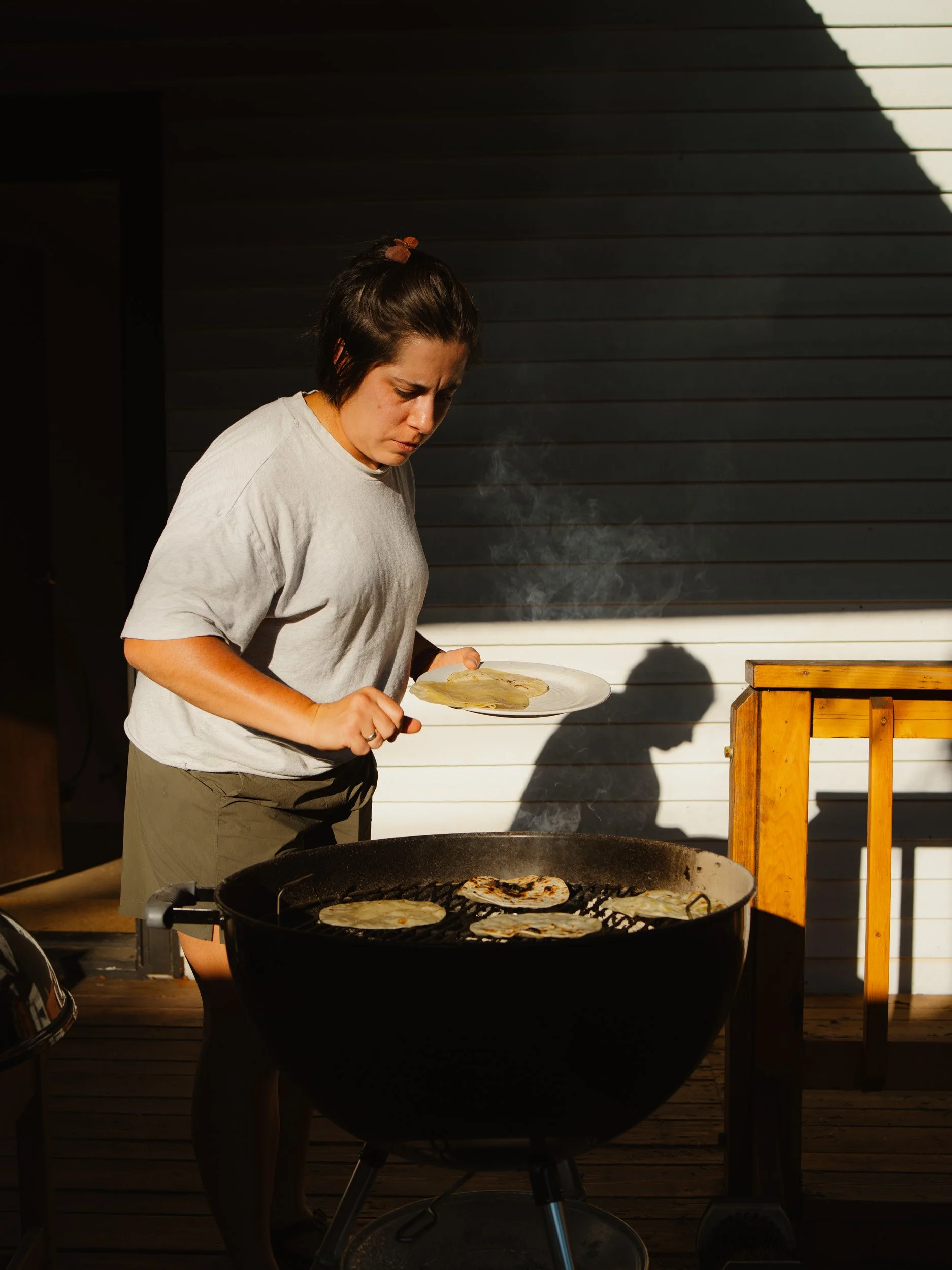 A woman is grilling tortillas on a barbecue grill, holding a plate with more tortillas while cooking outdoors.