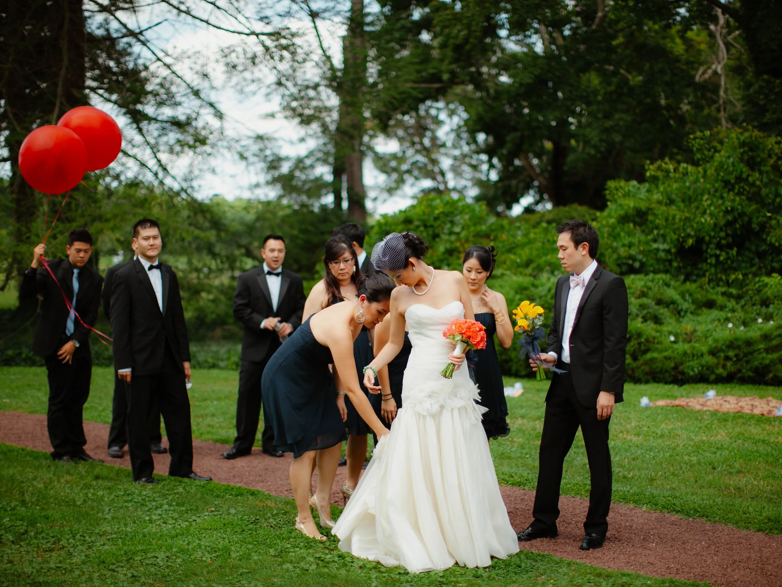 A bride in a white wedding dress standing on a grassy outdoor path holding a bouquet, surrounded by friends and family in formal attire, some holding flowers or balloons, with green trees in the background.