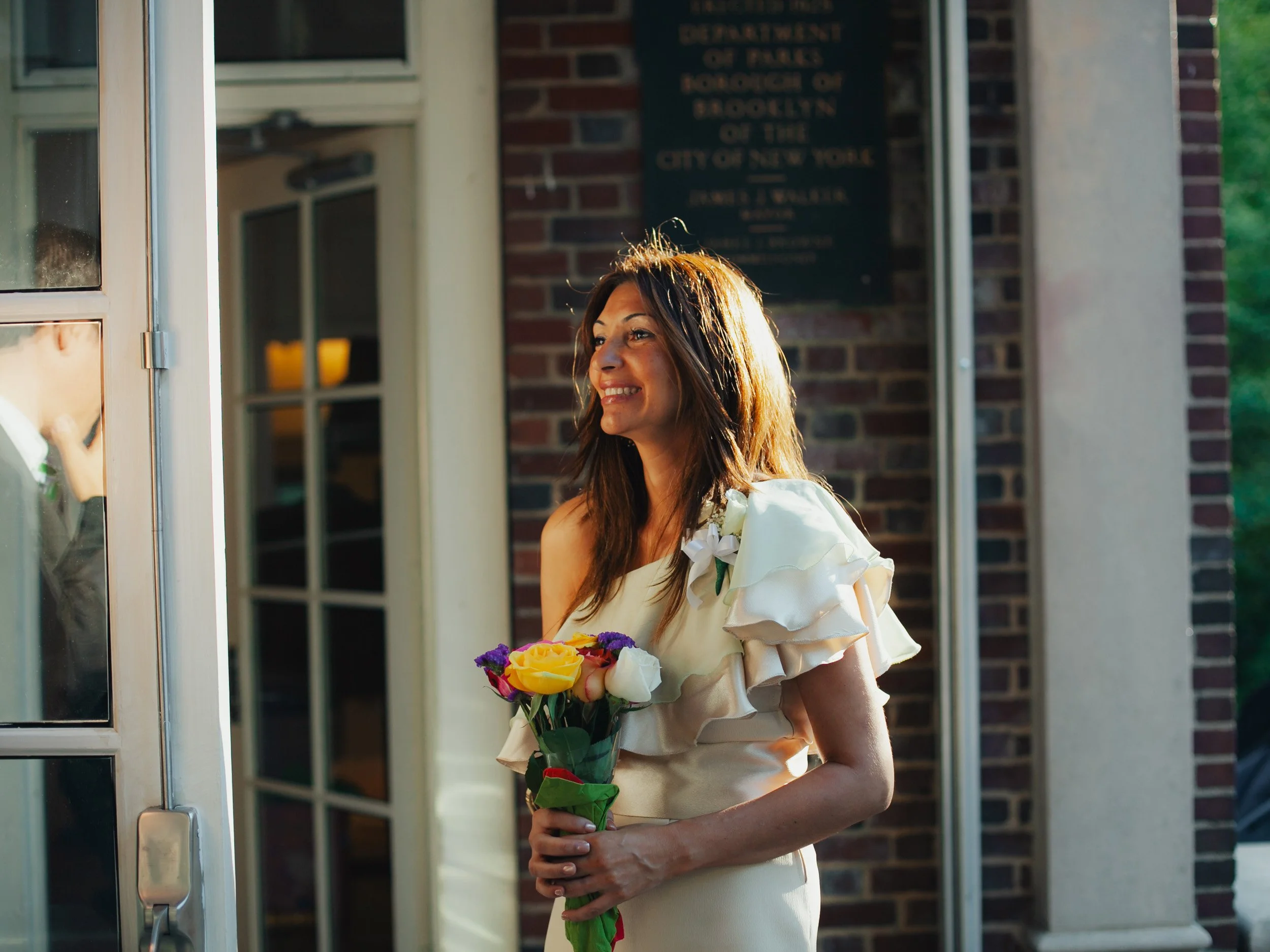 A woman in a cream-colored dress holding a bouquet of yellow, white, and purple flowers stands outside a building with a brick wall background, smiling and looking to the side.