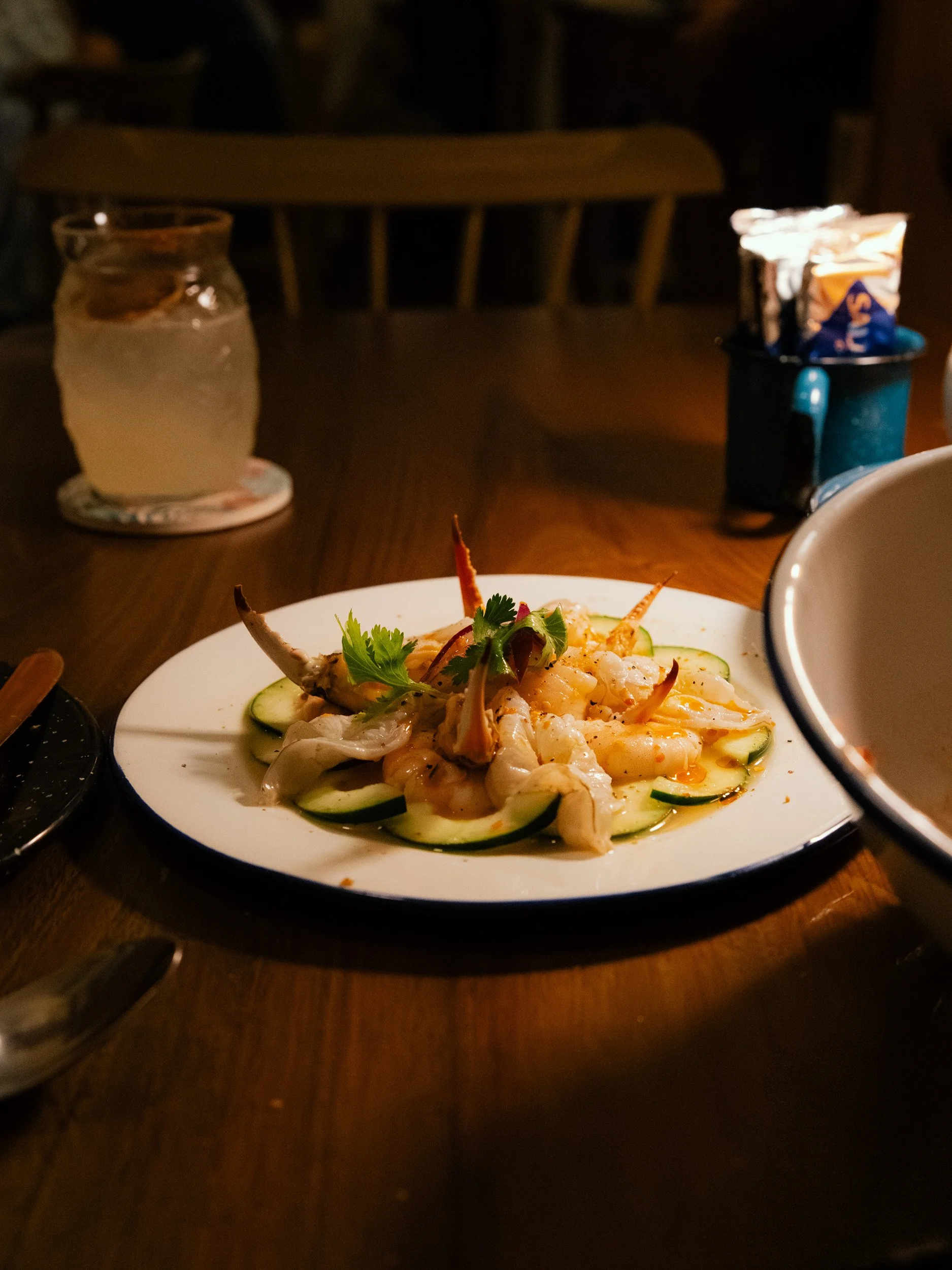 Seafood dish on a white plate with sliced cucumbers and cilantro garnishes, placed on a wooden table with water in a jar and napkins in a mug in the background.