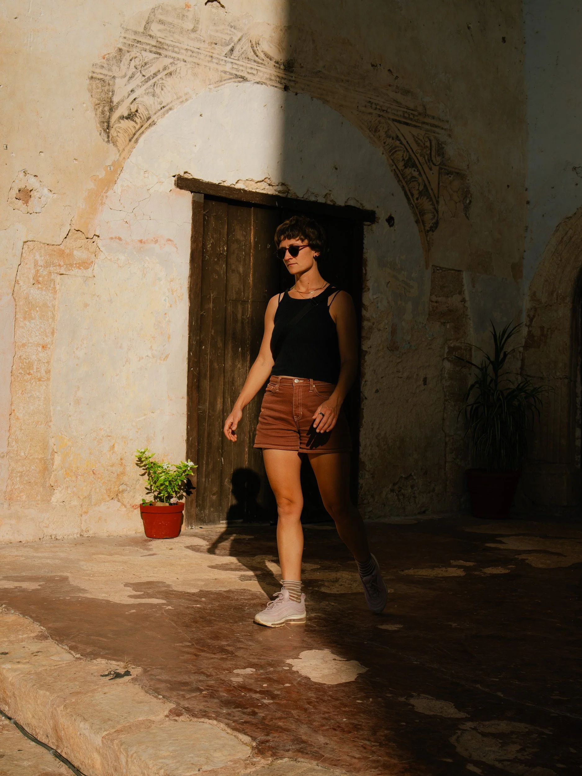 A young woman with sunglasses, a black sleeveless top, orange shorts, and sneakers walking in an ancient stone courtyard with aged walls and potted plants.