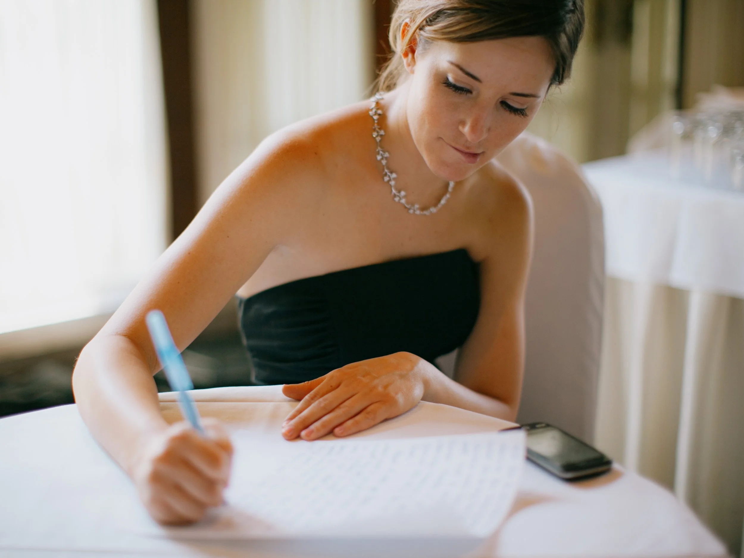 A woman in a black strapless dress sits at a table, writing in a notebook with a pen. She has short brown hair, is wearing a necklace, and has a focused expression. A smartphone is placed on the table beside her.