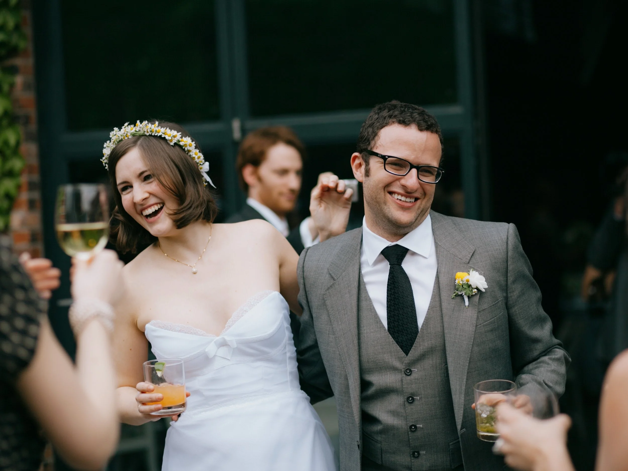 A group of wedding guests smiling and chatting at an outdoor reception, with a bride wearing a flower crown and a groom in a gray suit with a boutonniere, holding drinks.