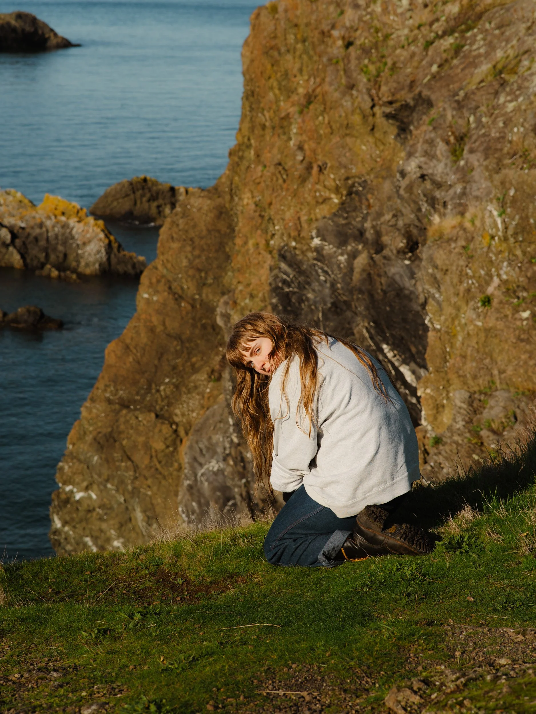 A woman kneeling on grass near rocky cliffs with ocean in the background.