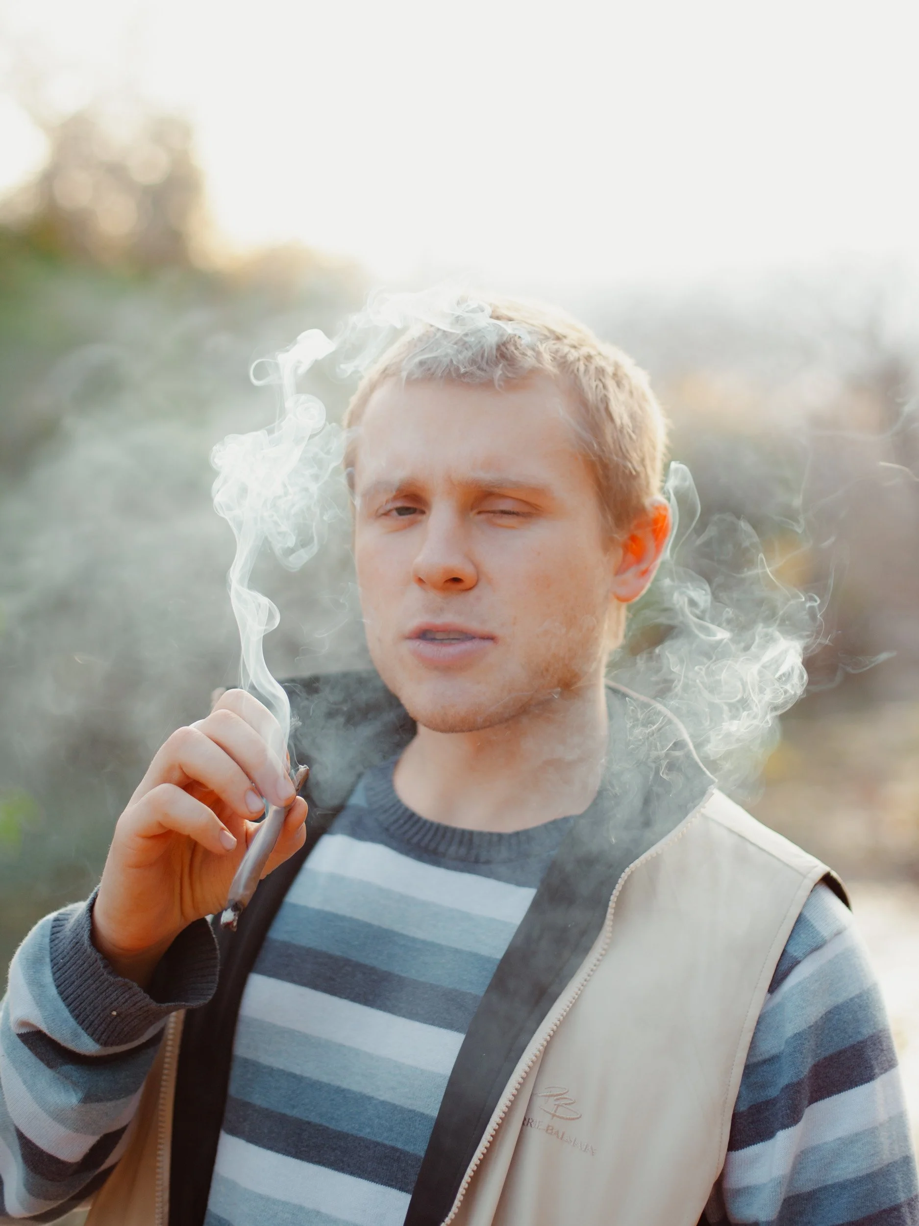 Young man outdoors smoking a cigarette, winking, during daytime with blurred foliage background.