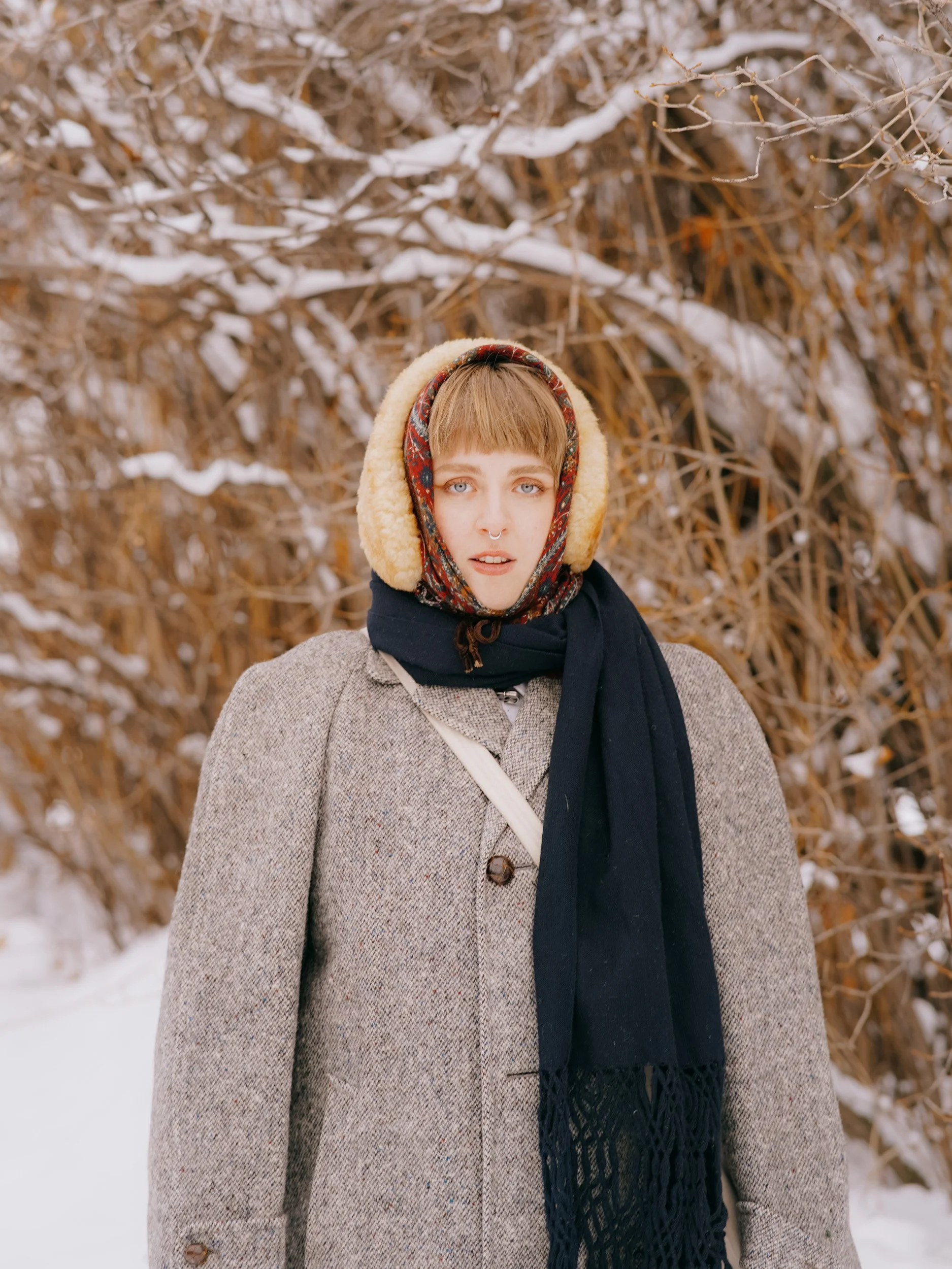 A woman wearing a grey coat, black scarf, red patterned scarf, and yellow earmuffs standing outdoors in front of snow-covered brown bushes.