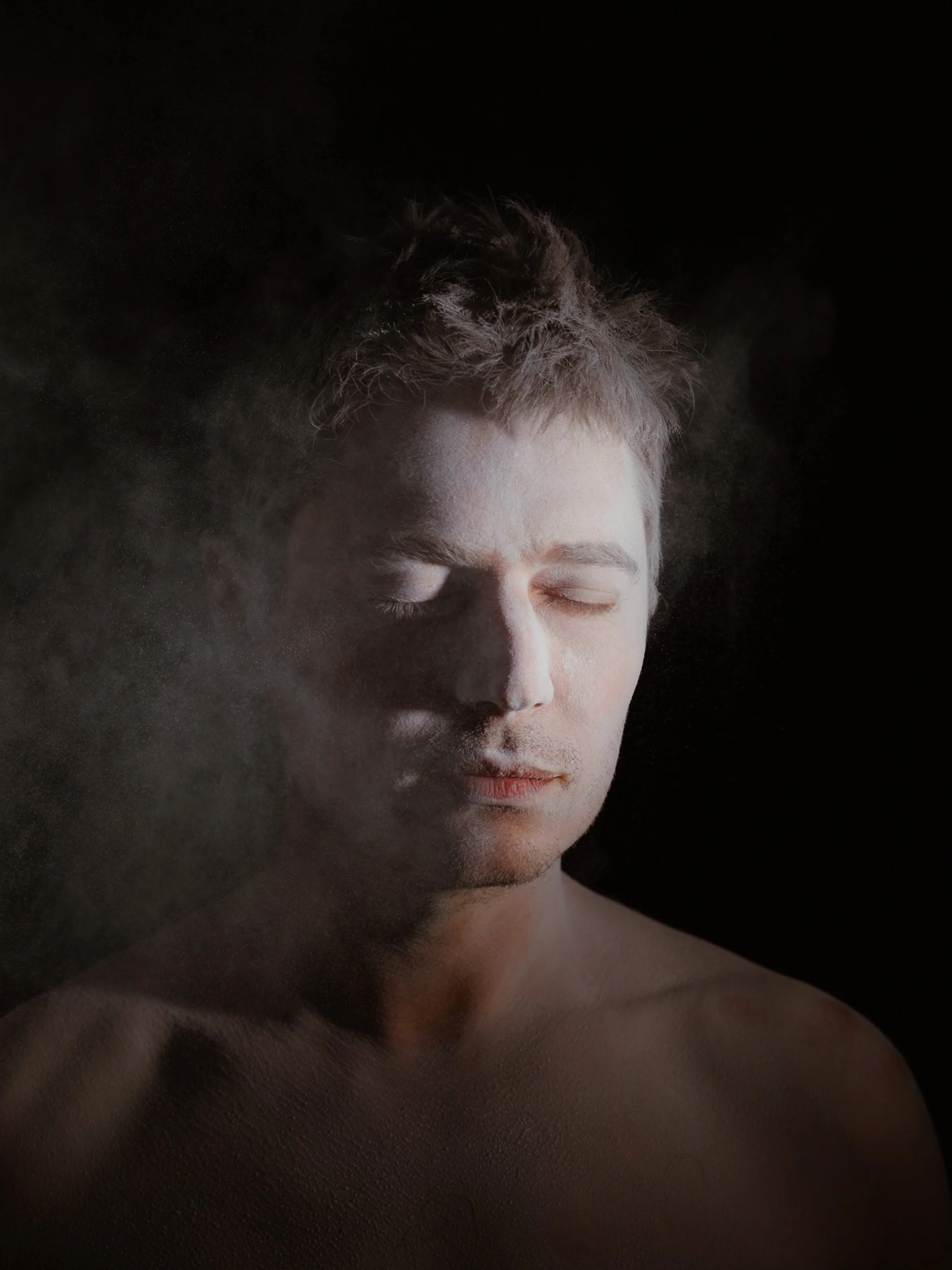 Close-up of a young man with closed eyes and a neutral expression, with light cloud or mist around his face and shoulder, against a dark background.