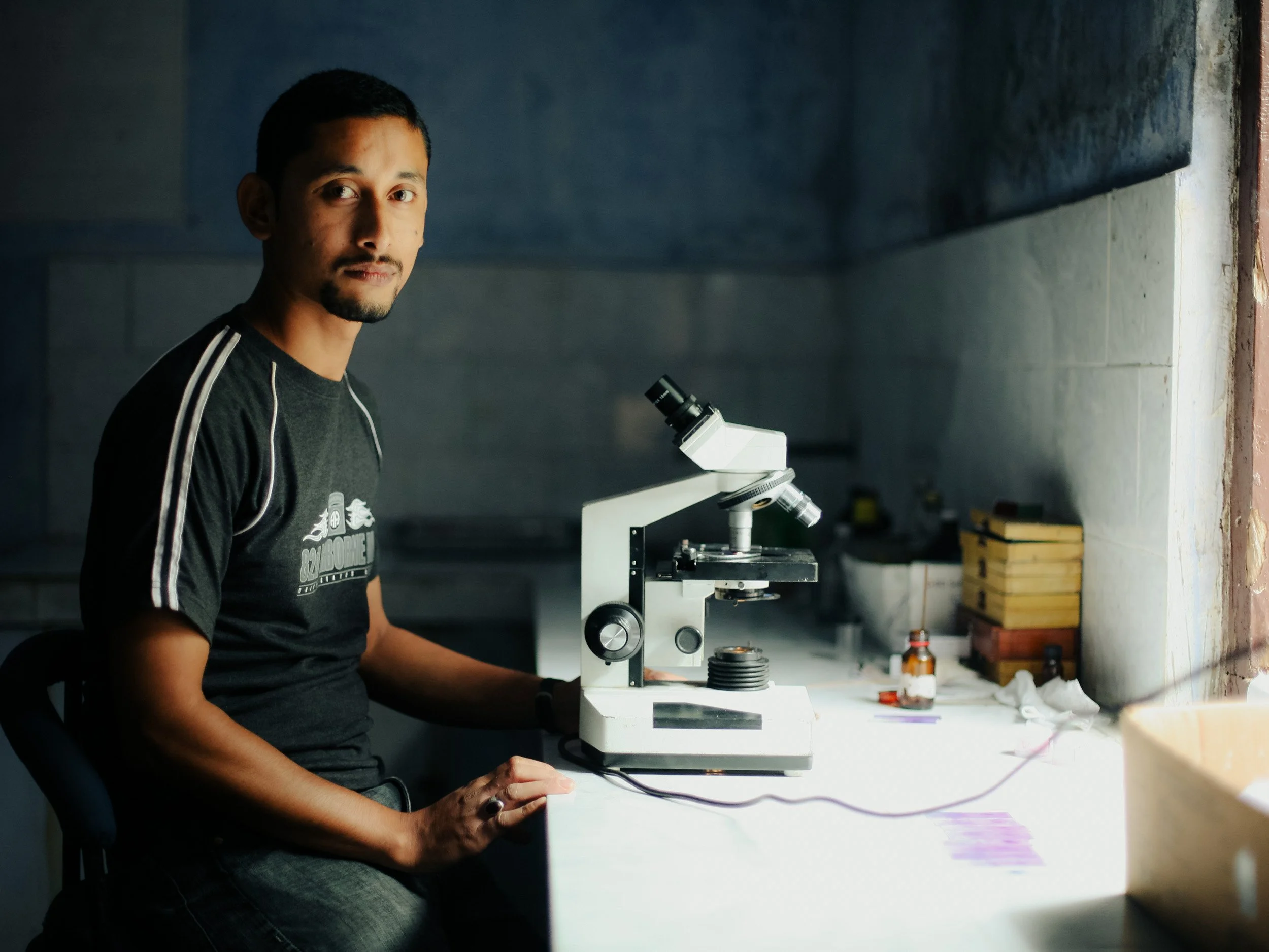 A man sitting at a laboratory bench with a microscope, in a slightly dimly lit room, with various laboratory supplies in the background.