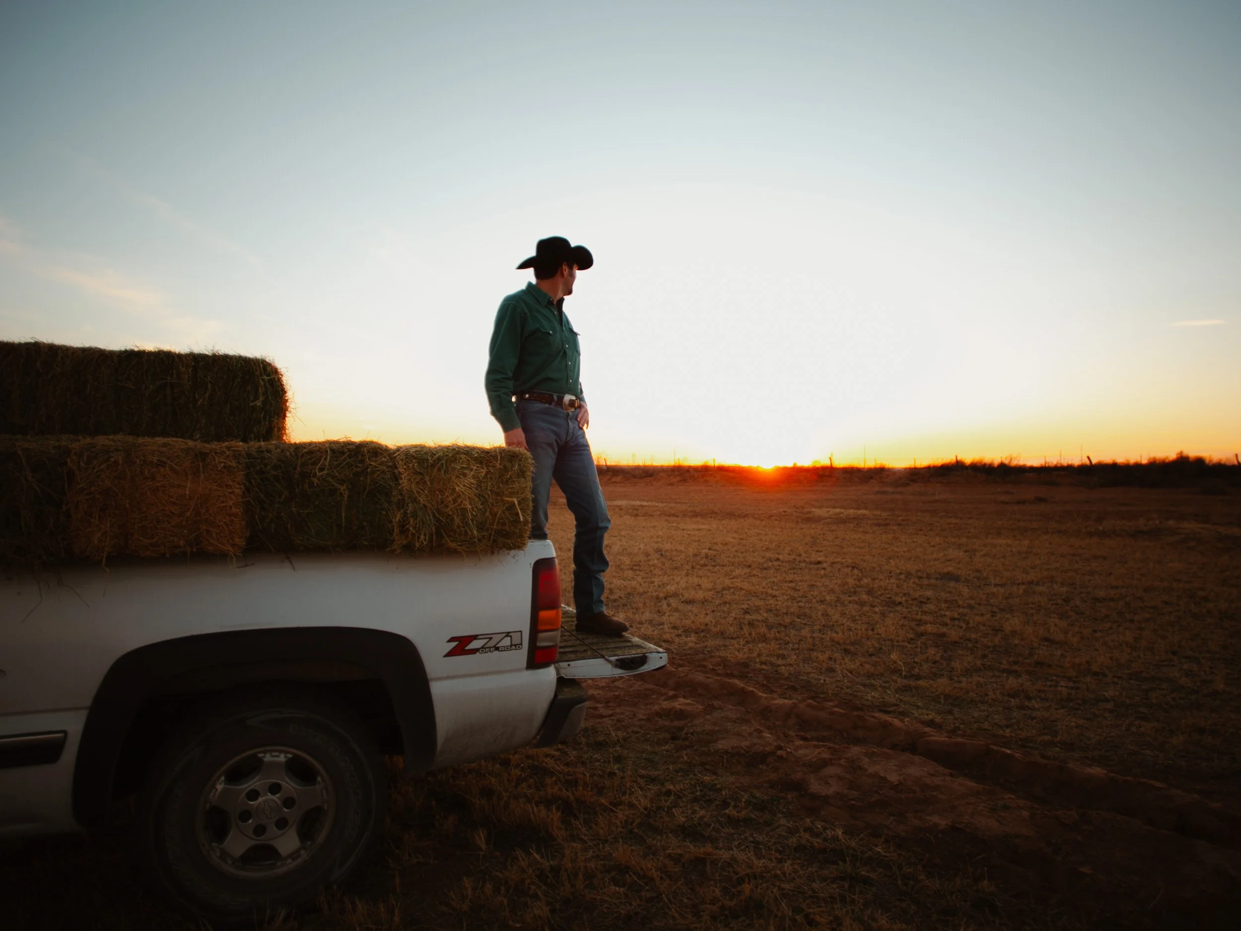 A person in a cowboy hat standing in the back of a pickup truck loaded with hay, watching the sunset in a rural field.