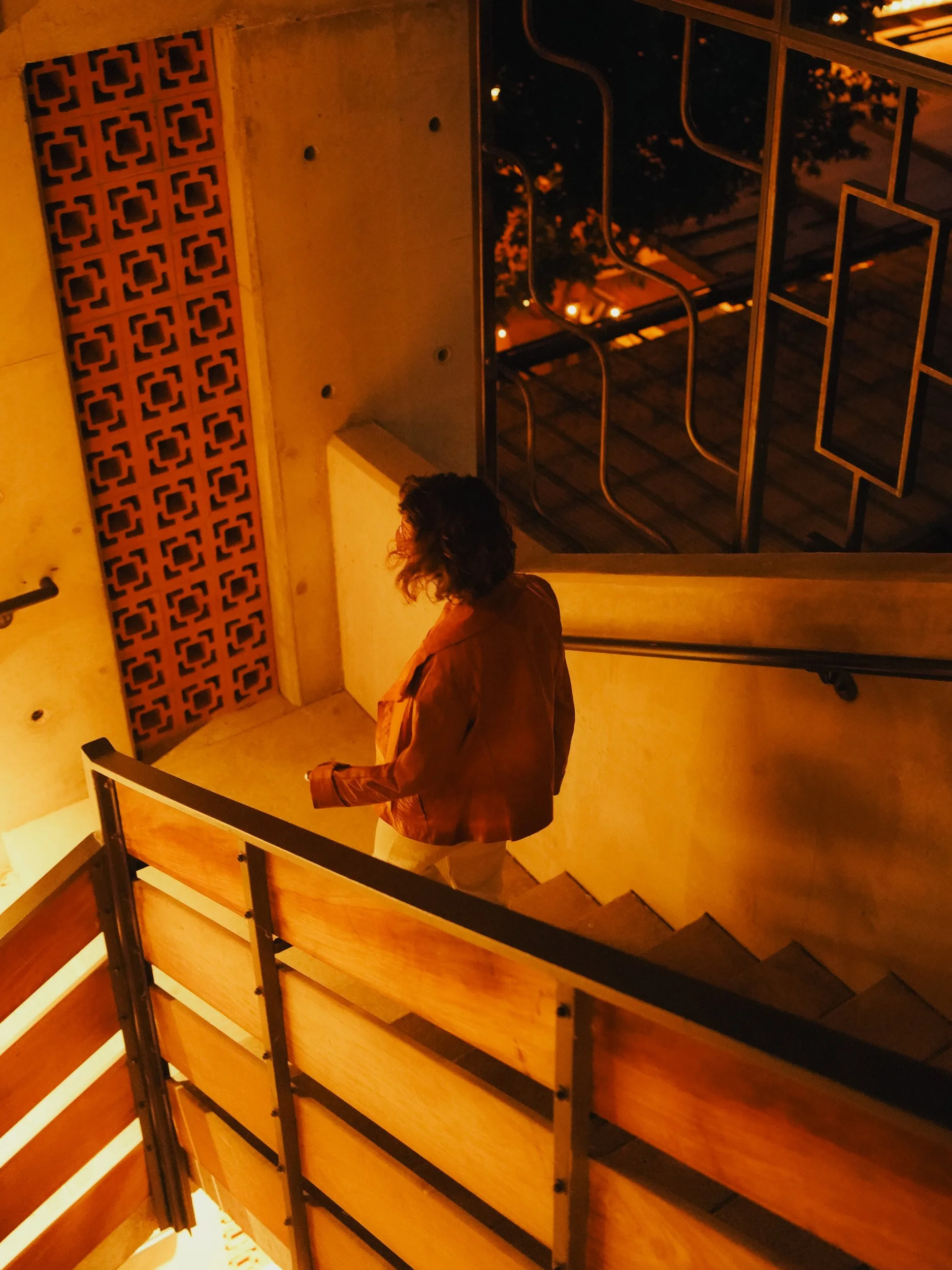 Person with curly hair wearing a brown jacket walking down stairs in a warmly lit indoor space at night.
