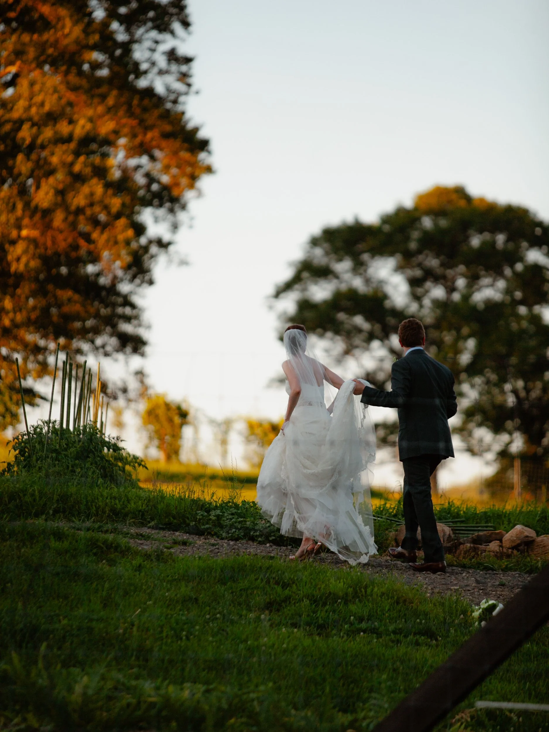 A bride and groom walking outdoors on a grassy path during sunset, with large trees in the background.