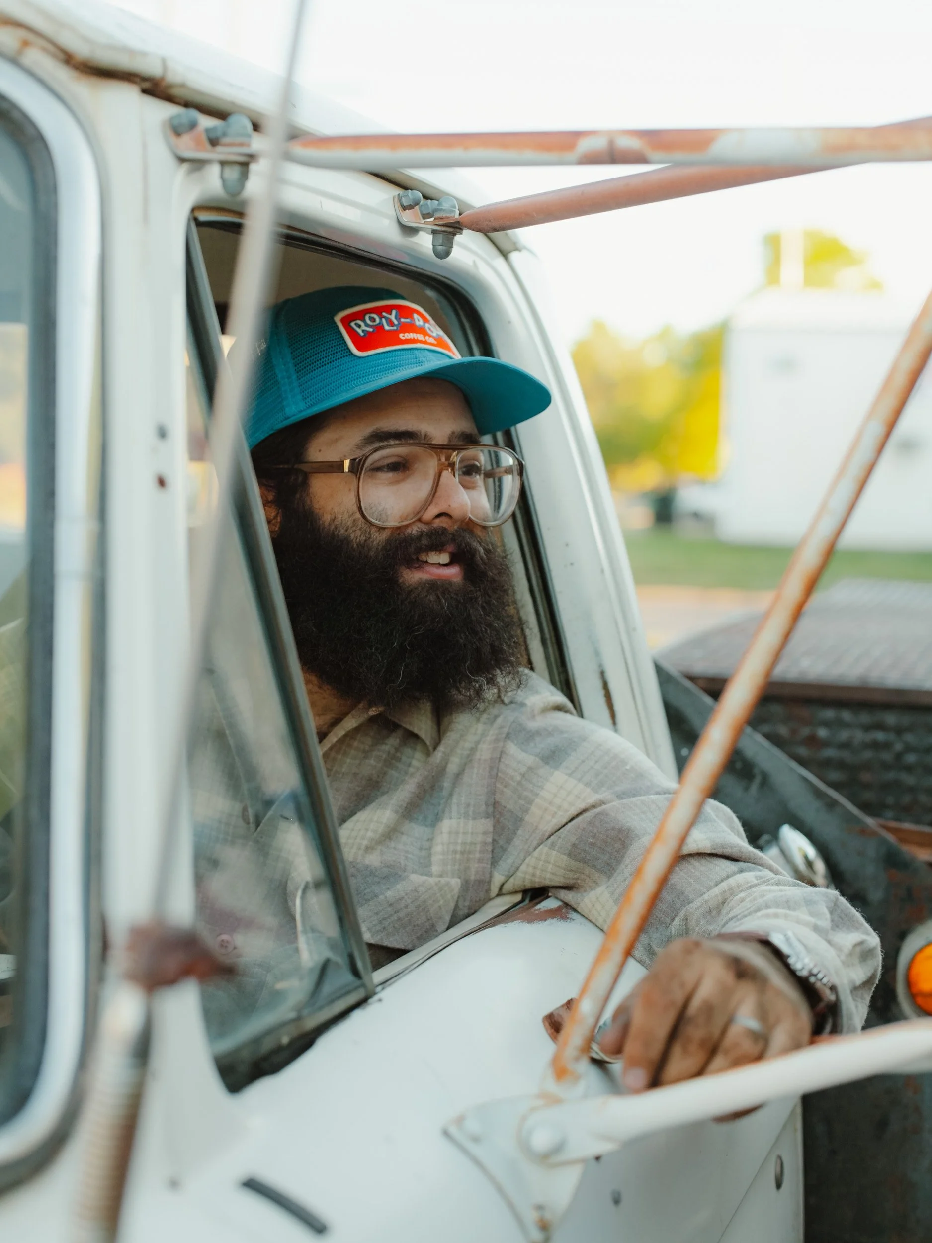A man with a beard, glasses, wearing a plaid shirt and a blue trucker hat with a red and white logo, sitting in the driver's seat of a vintage white truck, looking out of the window.