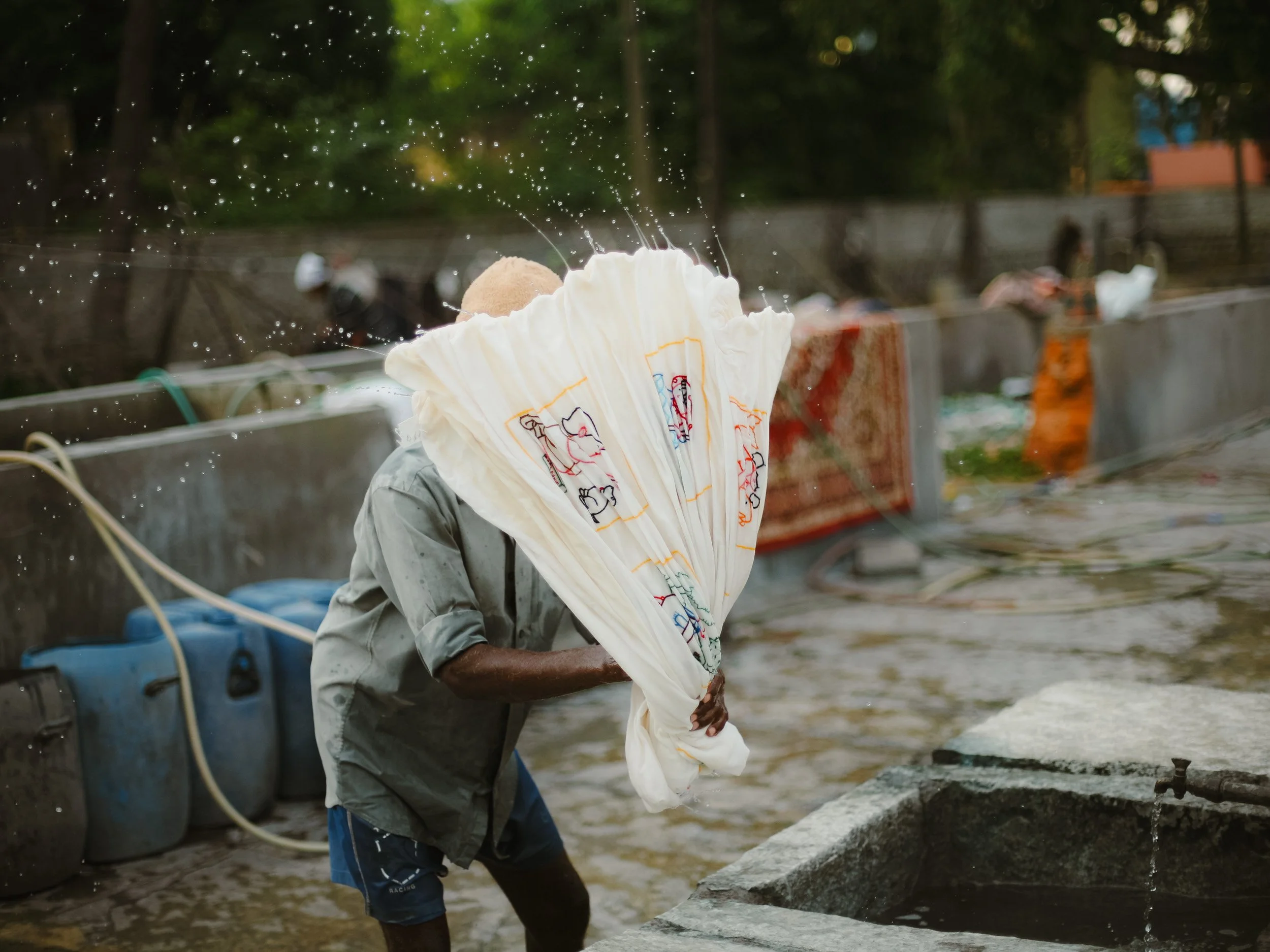 Person rinsing or washing a large, white cloth with colorful embroidery at a water well, water splashing from the cloth. The individual is outdoors in a setting with containers and trees in the background.