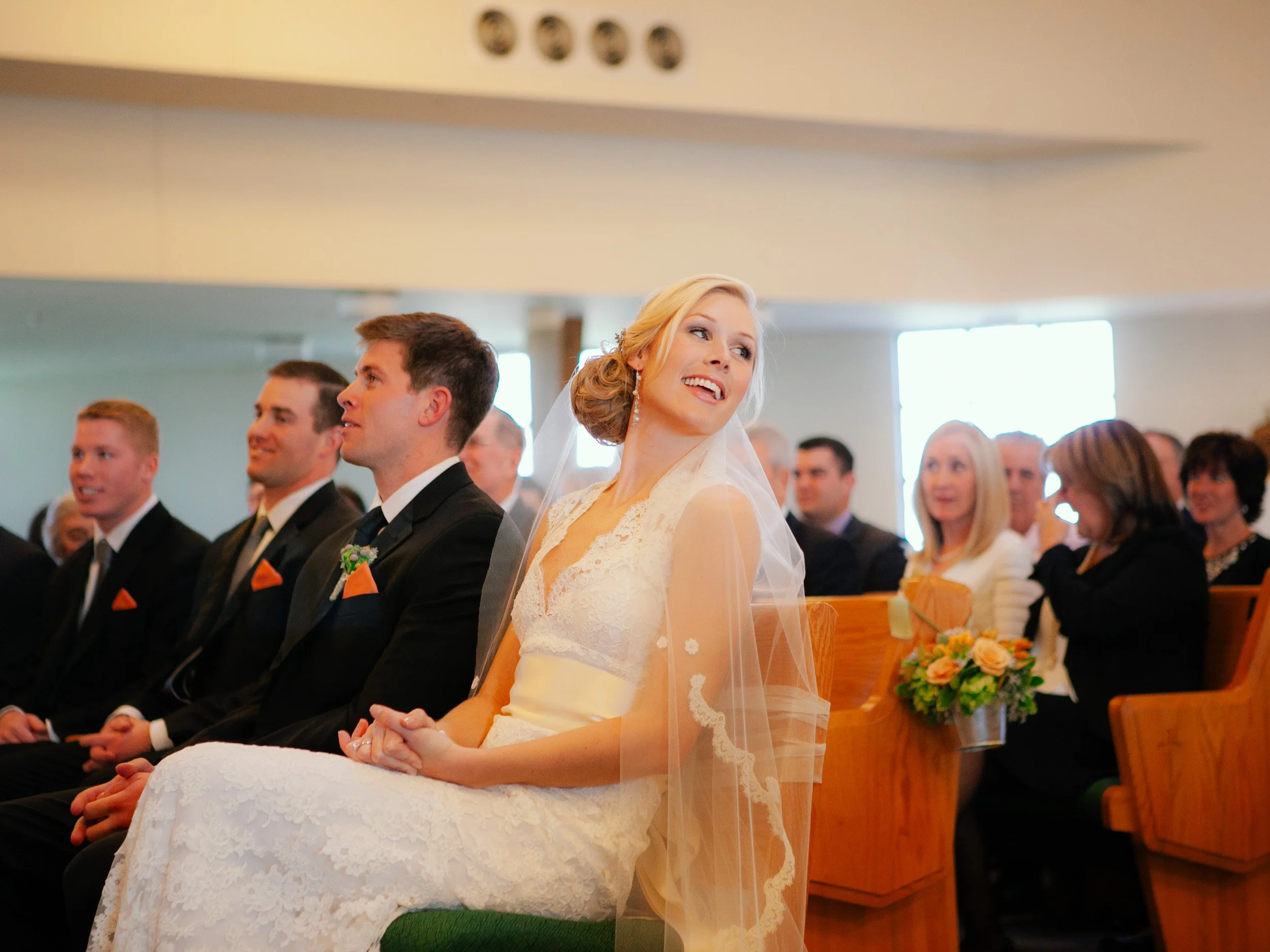 Bride smiling during wedding ceremony, seated among guests in church or chapel.
