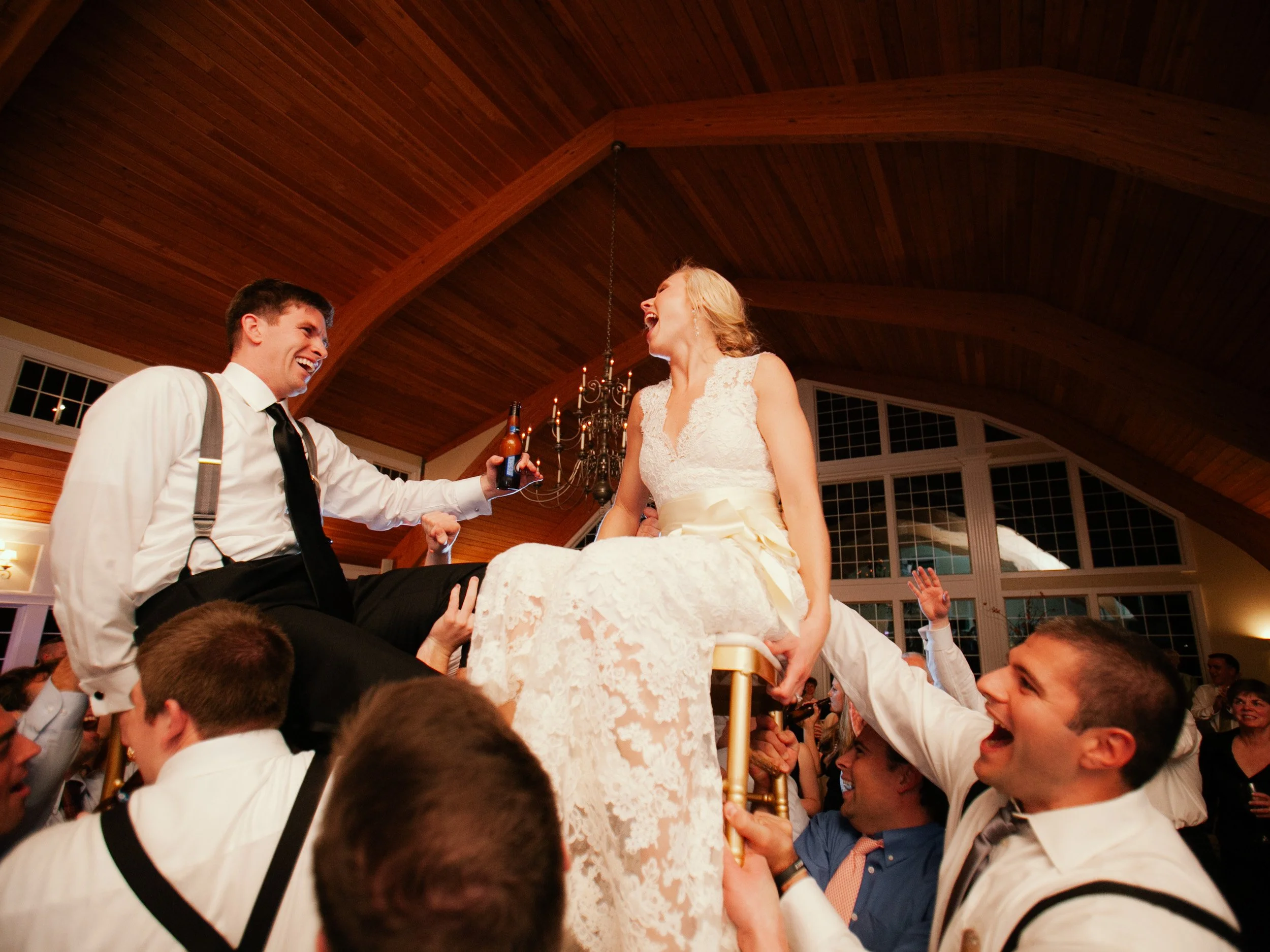 People celebrating a wedding, with a woman in a white lace wedding dress sitting on a chair held up by a man, surrounded by guests cheering and smiling, inside a wooden ceiling venue.