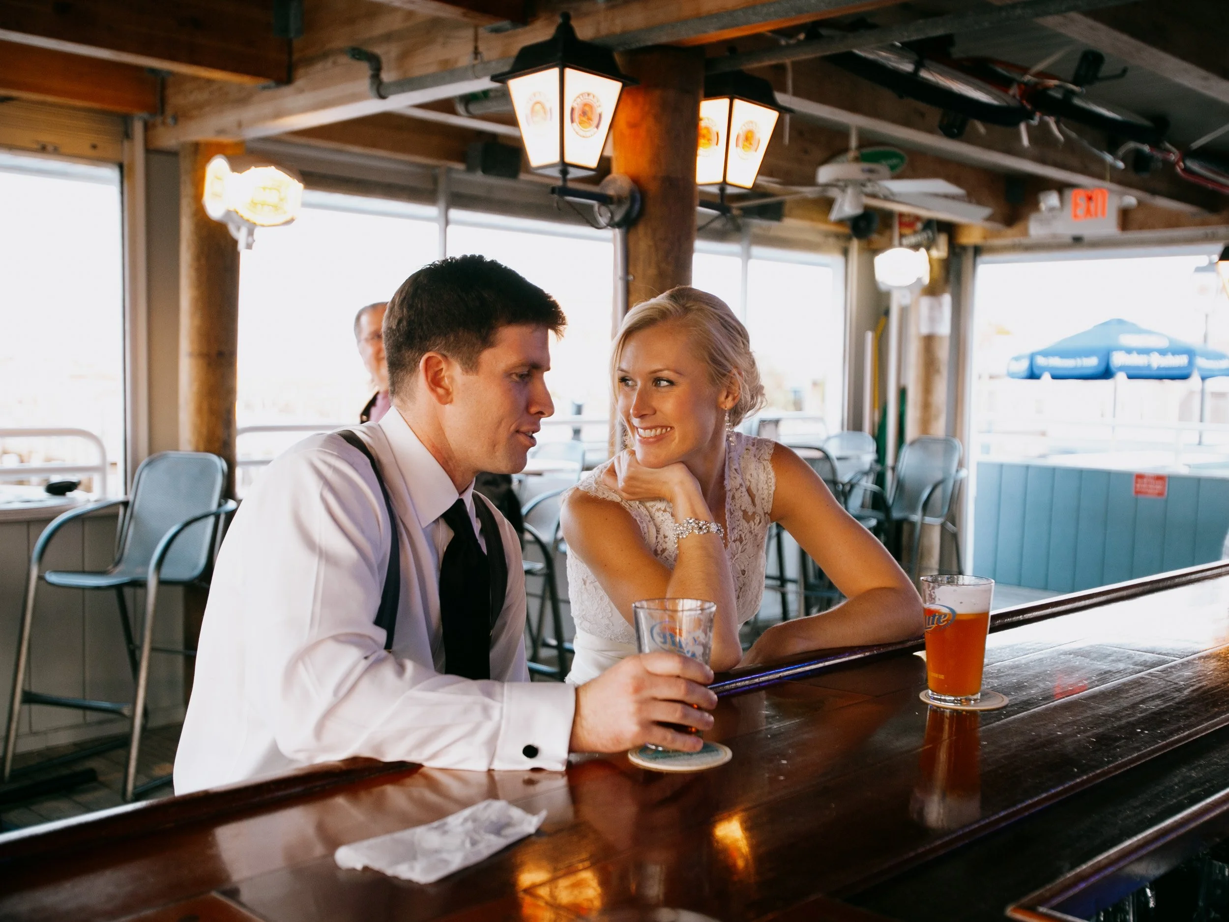 A man in a white shirt with suspenders and a woman in a white lace dress sit at a wooden bar, smiling and looking at each other. They each have a drink in front of them in a bar or restaurant with large windows and warm lighting.