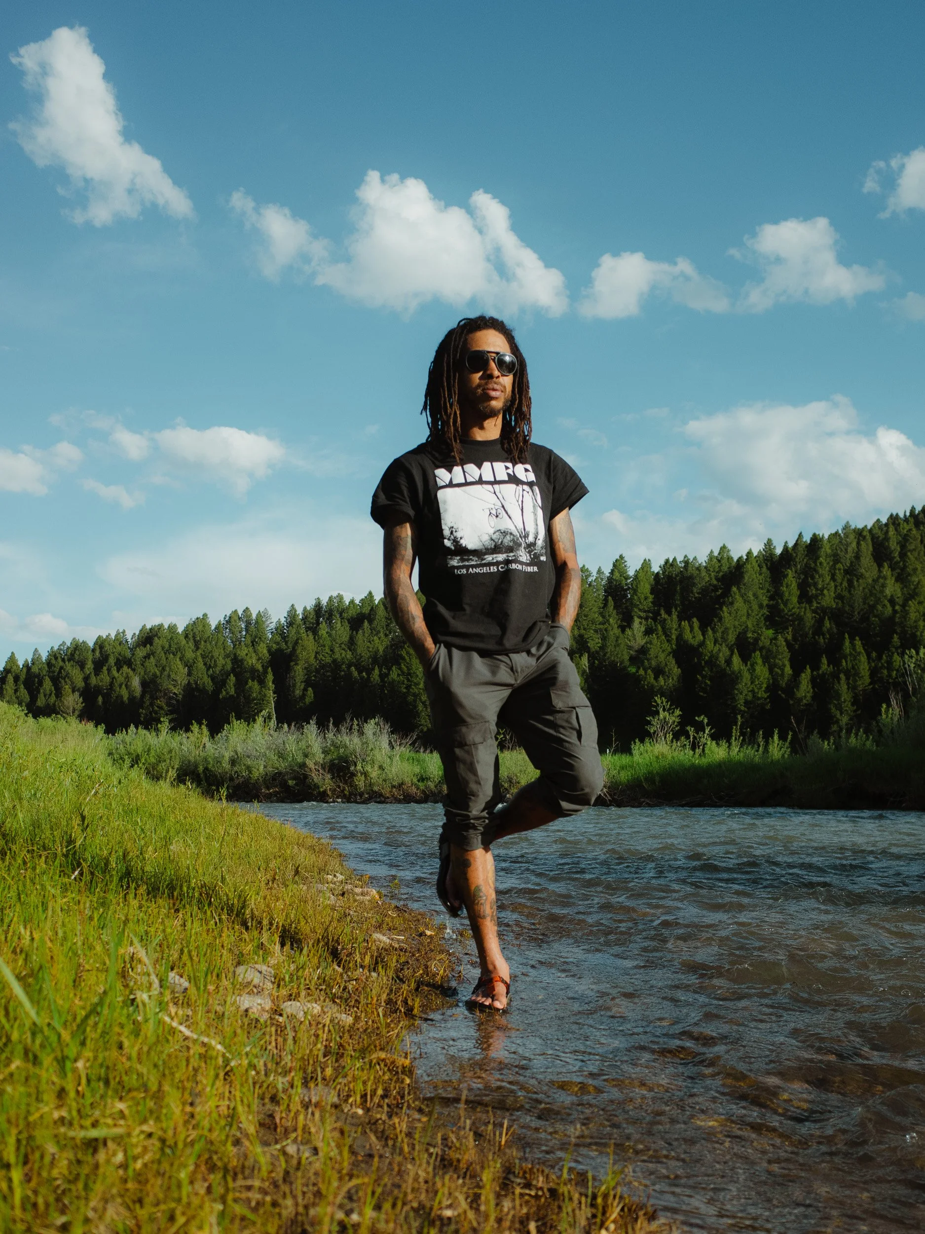 A man with dreadlocks and sunglasses standing in a shallow creek, surrounded by green grass and trees, with a blue sky and clouds above.