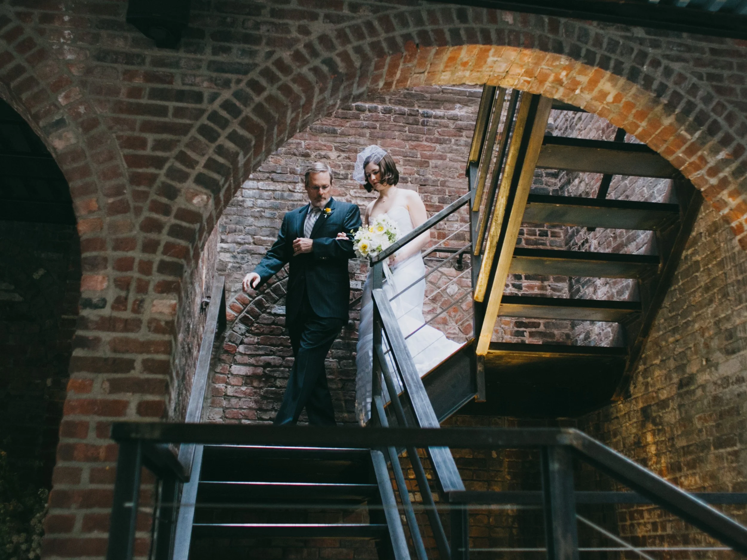 A bride in a white wedding dress holding a bouquet and an older man in a dark suit walking down a brick staircase inside an industrial building with brick archways.