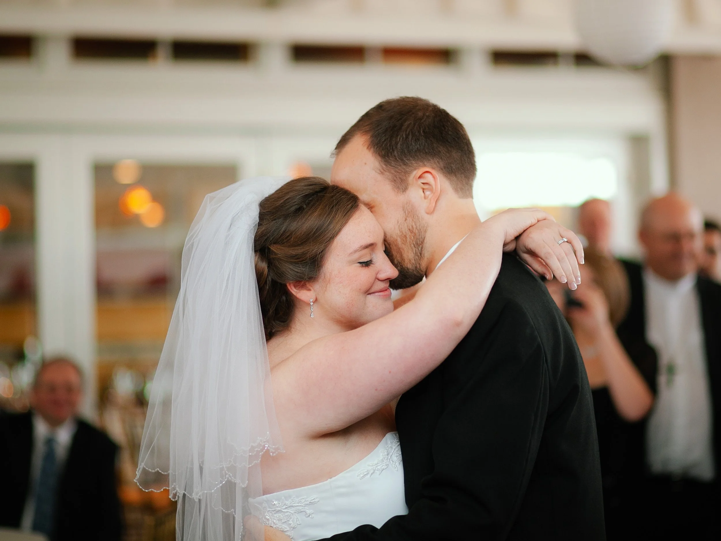 Bride and groom sharing a first dance at their wedding, embracing close with closed eyes, surrounded by wedding guests.