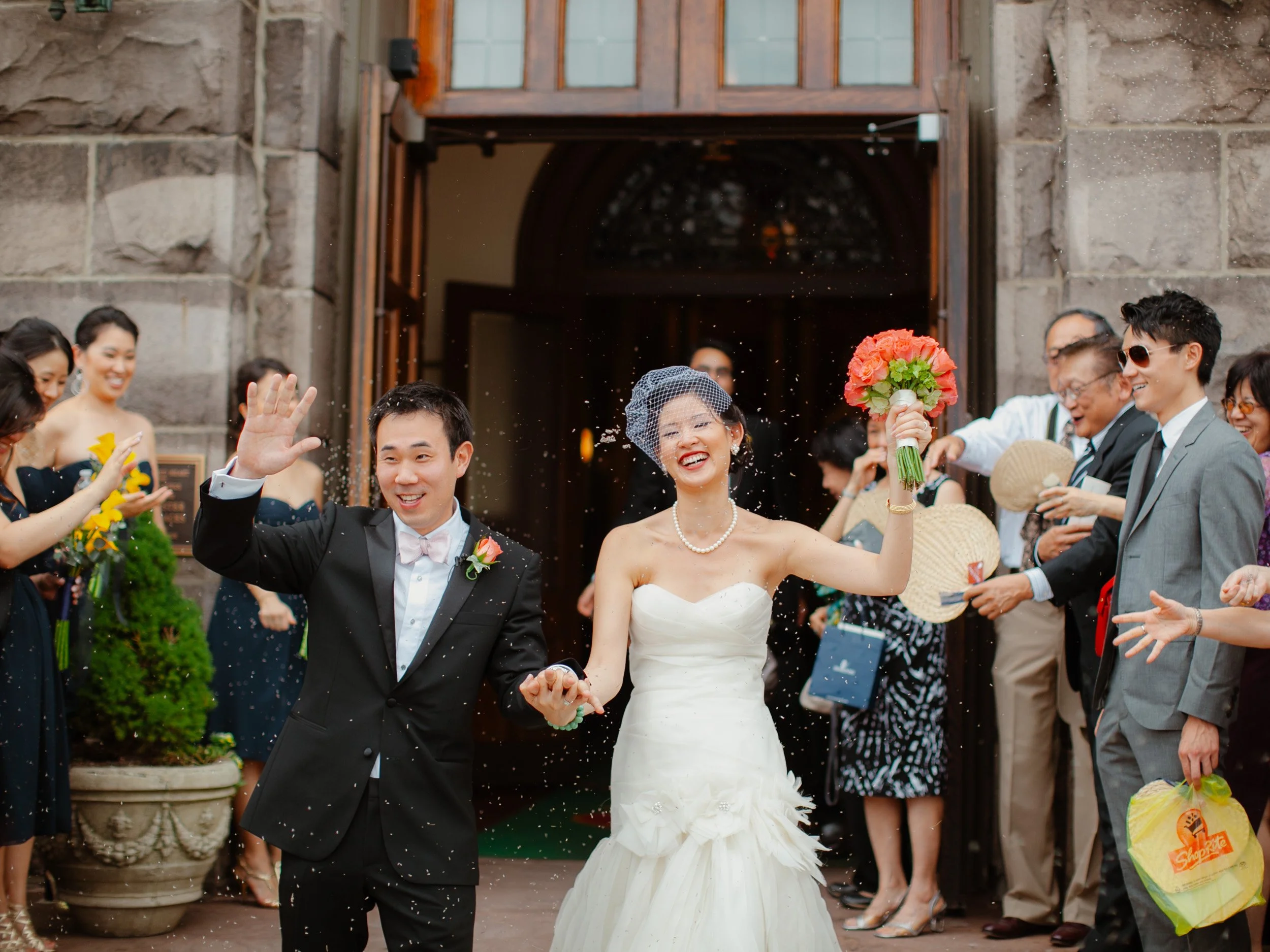 Bride and groom walking out of a building, smiling and holding hands, surrounded by smiling guests. The bride is wearing a strapless white wedding gown, pearls, and holding a bouquet of red flowers. The groom is in a black tuxedo with a pink bow tie 