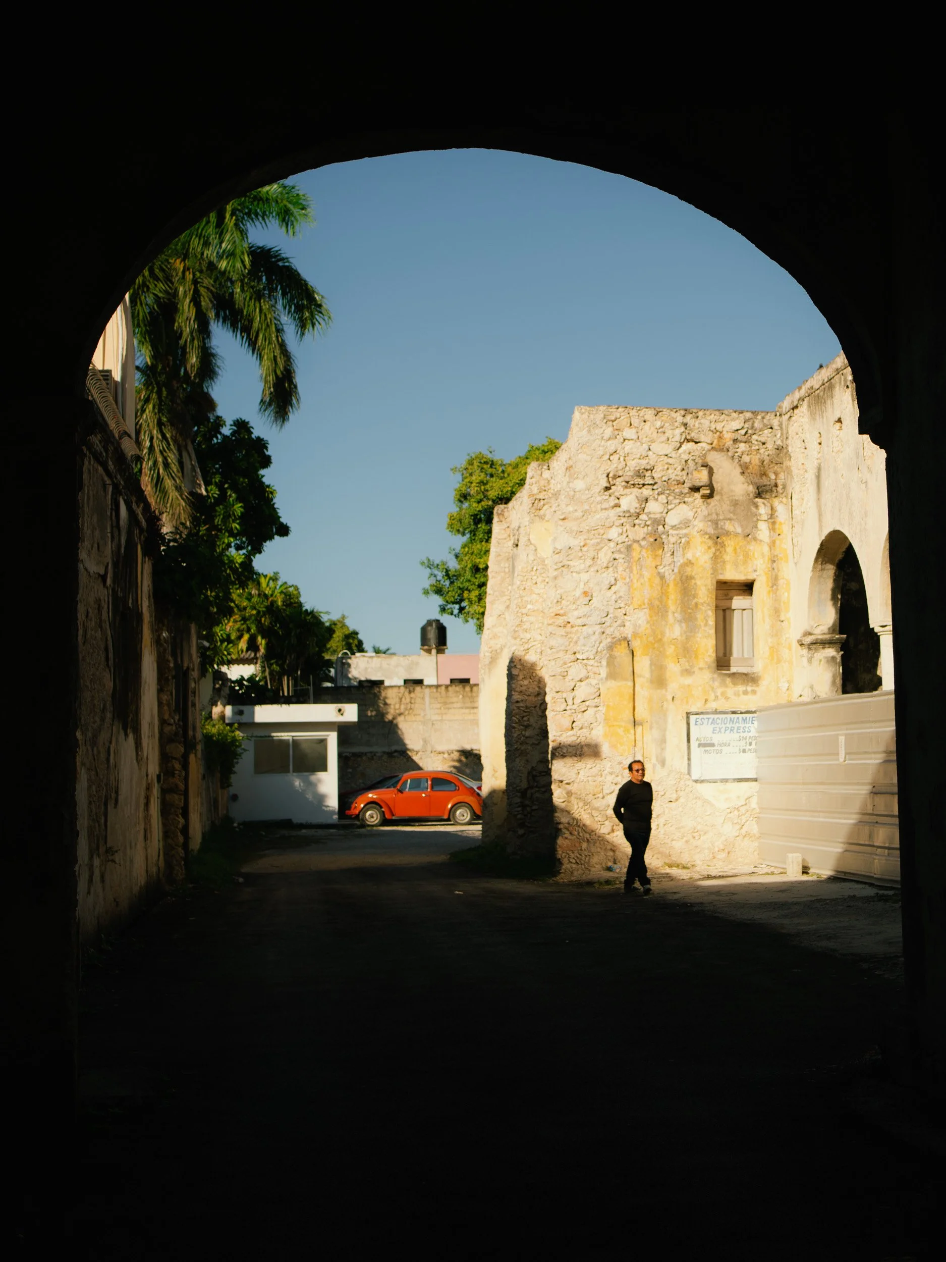 View of a street seen from under an arch, featuring an old stone building, a person walking, a red vintage car, and palm trees under a clear blue sky.