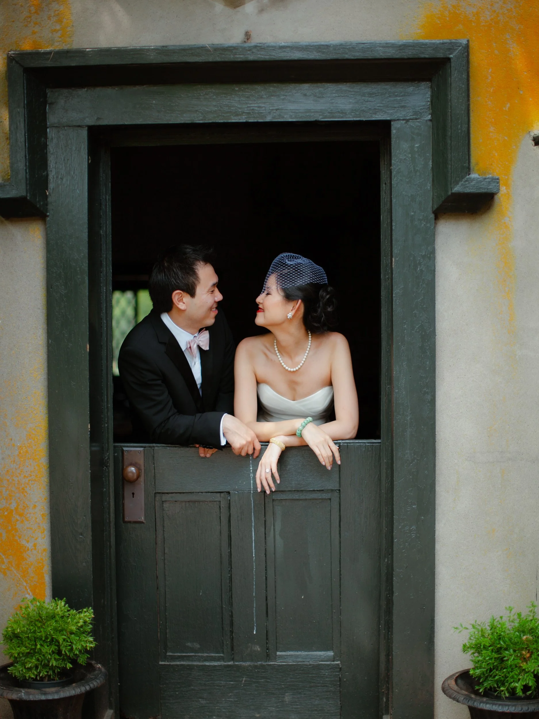A bride and groom leaning on a dark green door frame, sharing a joyful moment, inside a rustic setting.