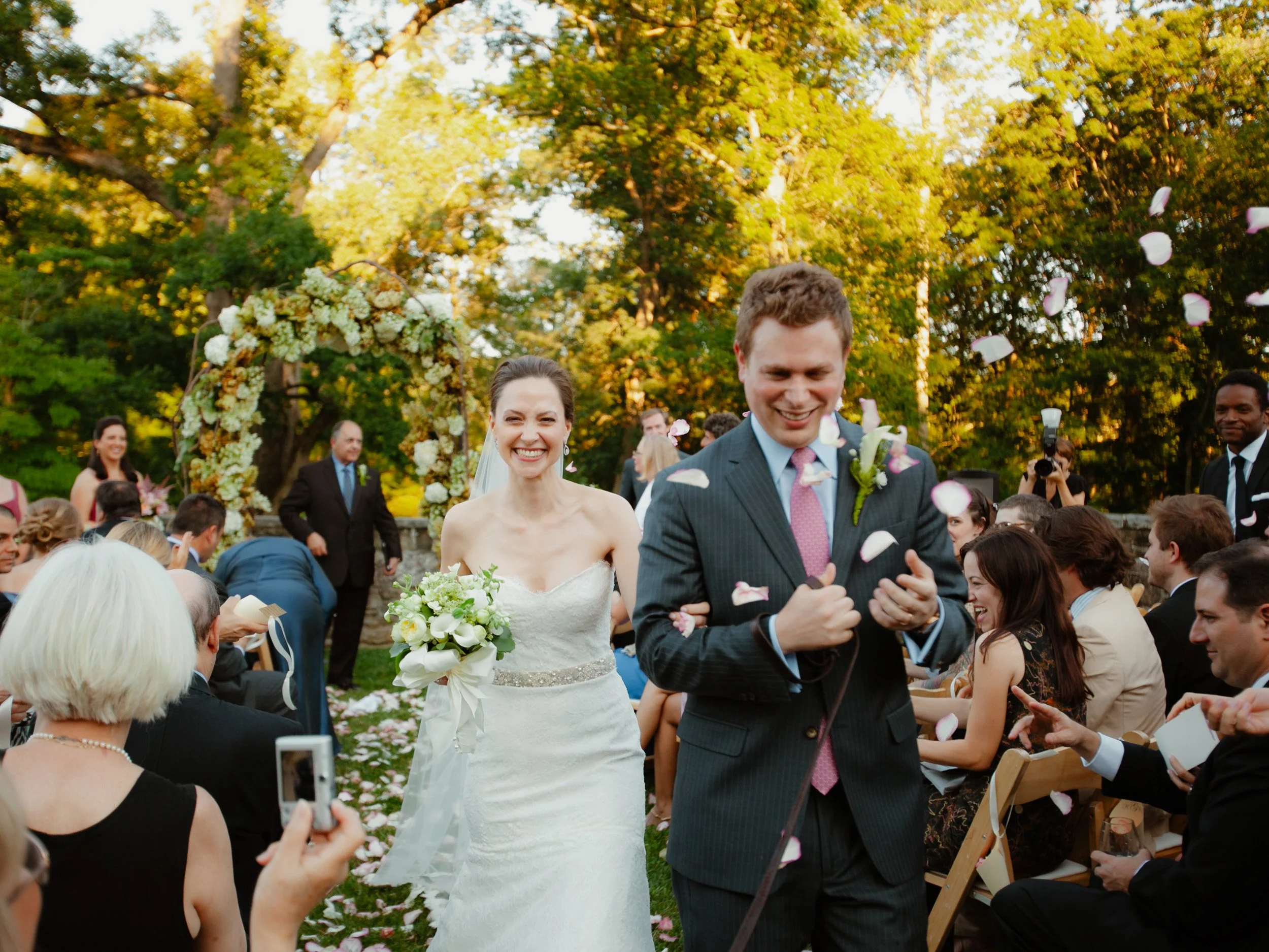 A wedding ceremony outdoors with a bride in a white strapless gown holding a bouquet and a groom in a suit with a pink tie, smiling as they walk down the aisle amid falling flower petals, surrounded by seated guests, a floral arch, and green trees.
