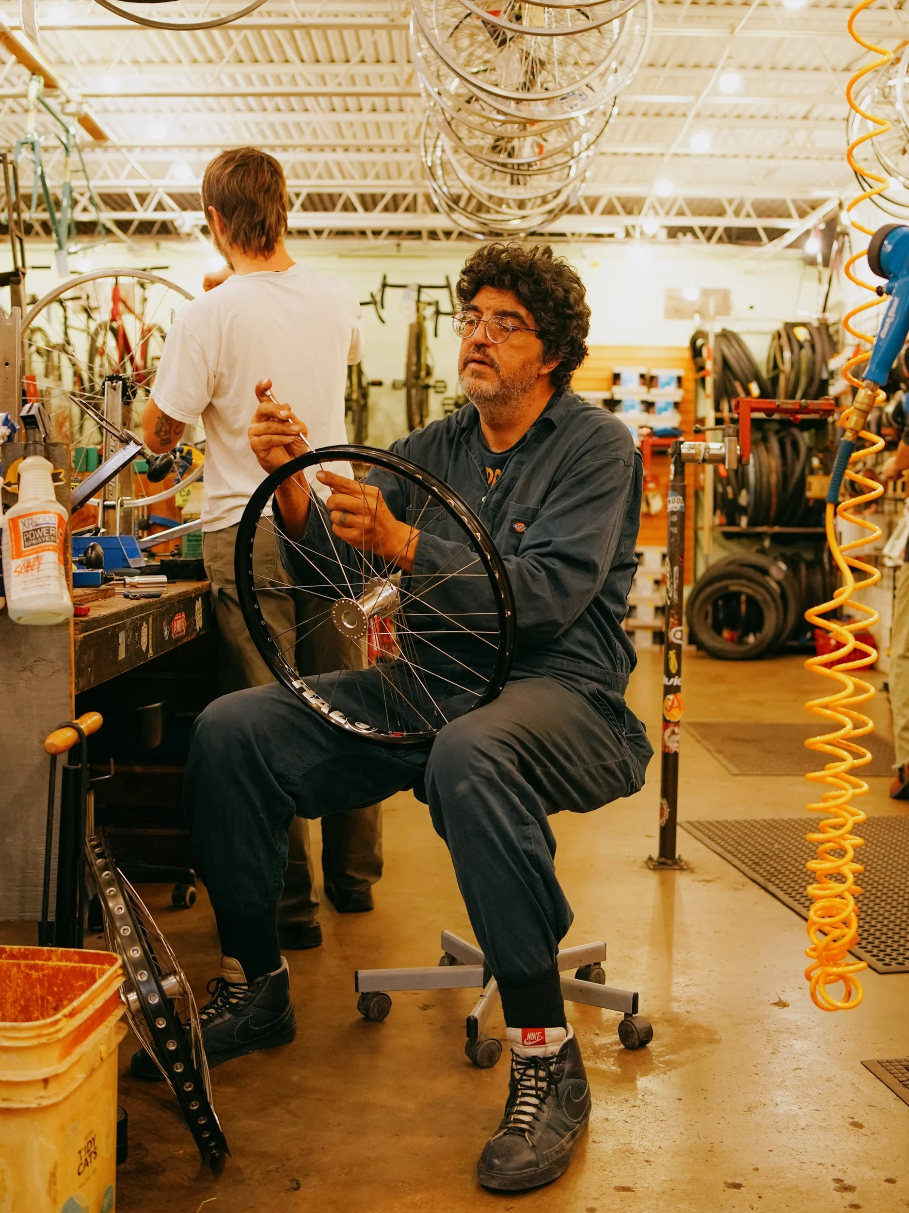 A man with curly hair and glasses working on a bicycle wheel inside a bike shop, with another person in the background.