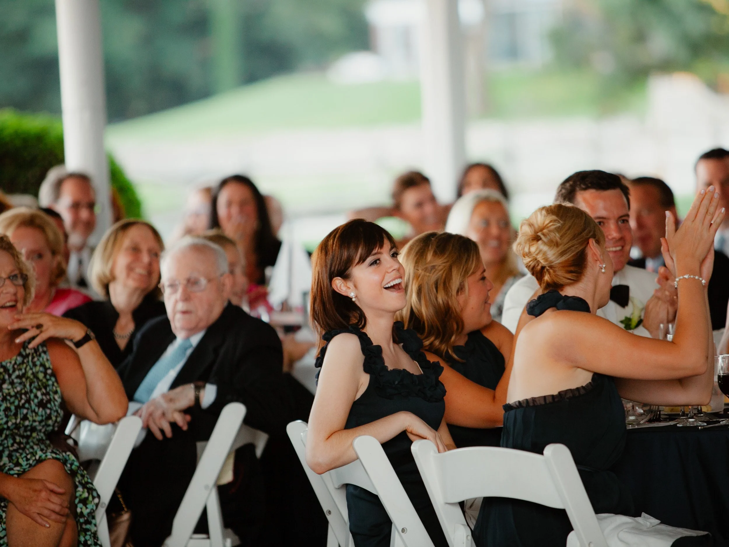 A group of people sitting at a long table at a wedding reception, laughing and smiling, with a woman's joyful expression in the center of the image.
