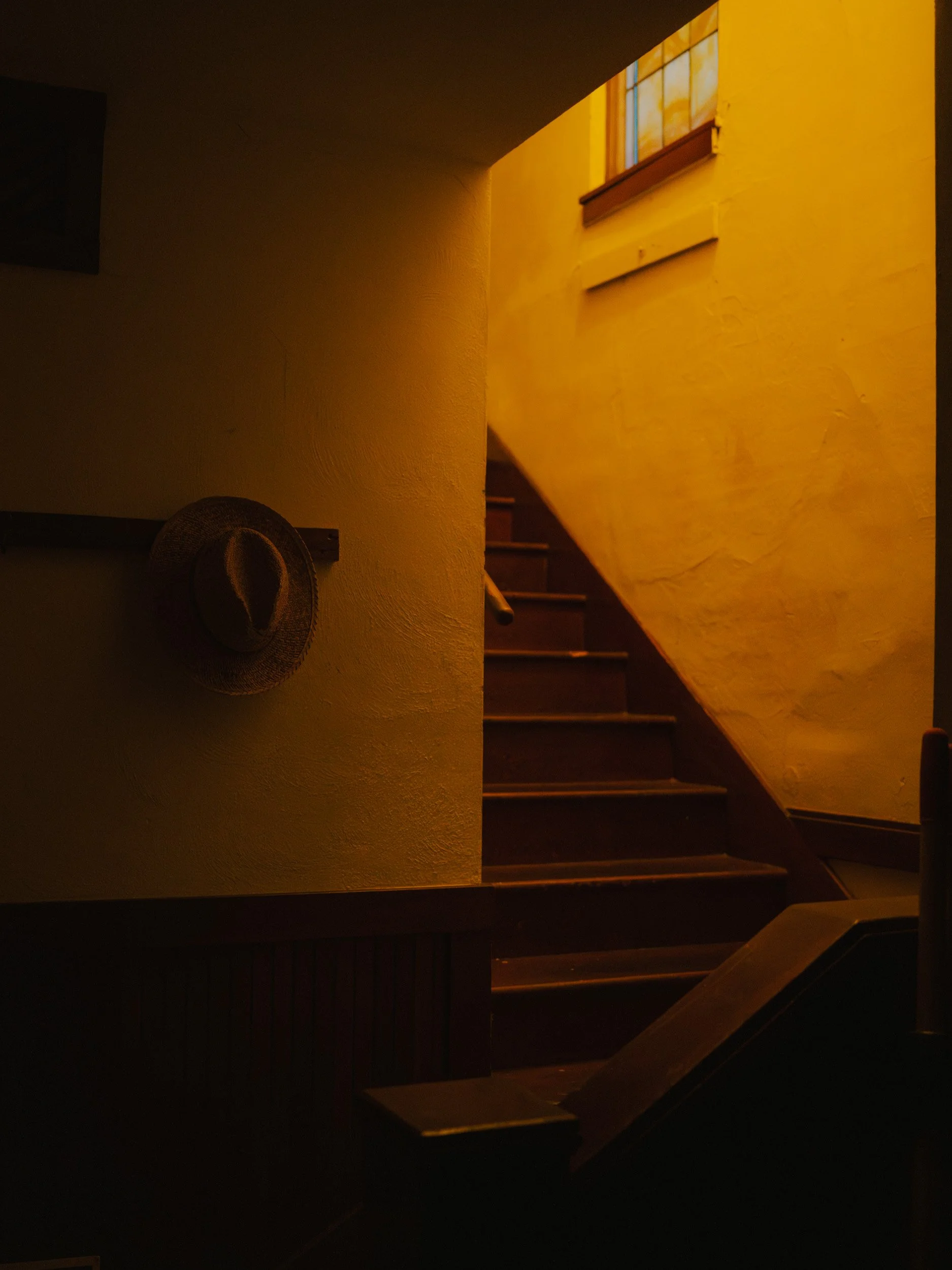 Interior of a dimly lit staircase with a yellow wall and a window at the top, a hat hanging on the wall.