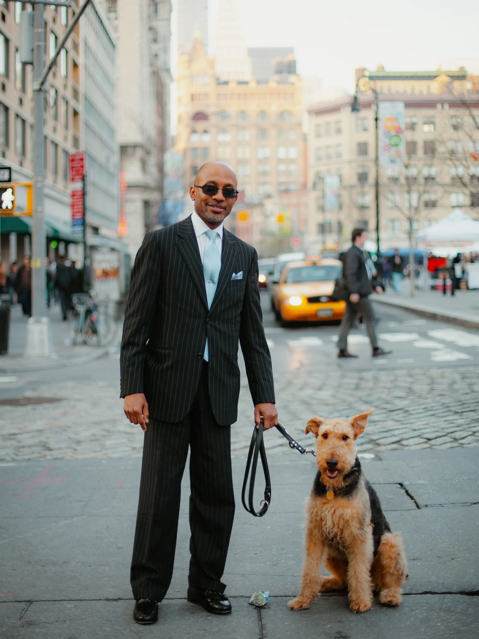 Man in a pinstripe suit and sunglasses standing with a dog on a city sidewalk in front of buildings and a yellow taxi.