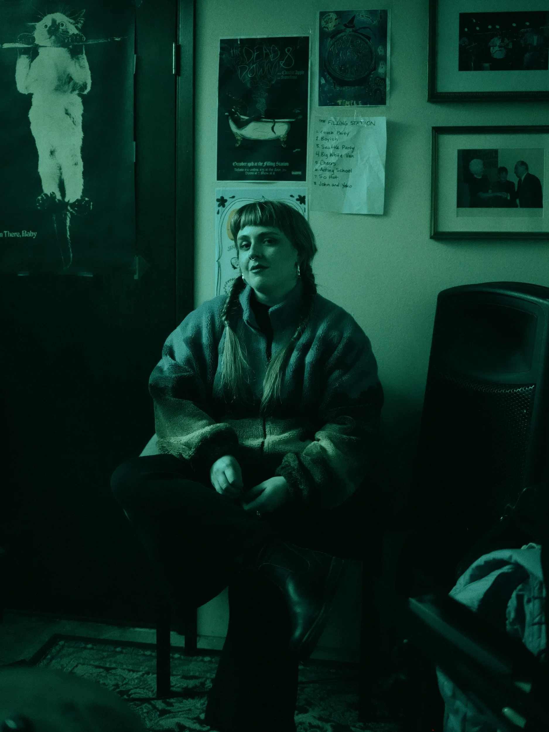 A woman with long hair and braid sitting on a chair, wearing a fleece jacket, in a room with various wall posters and pictures.