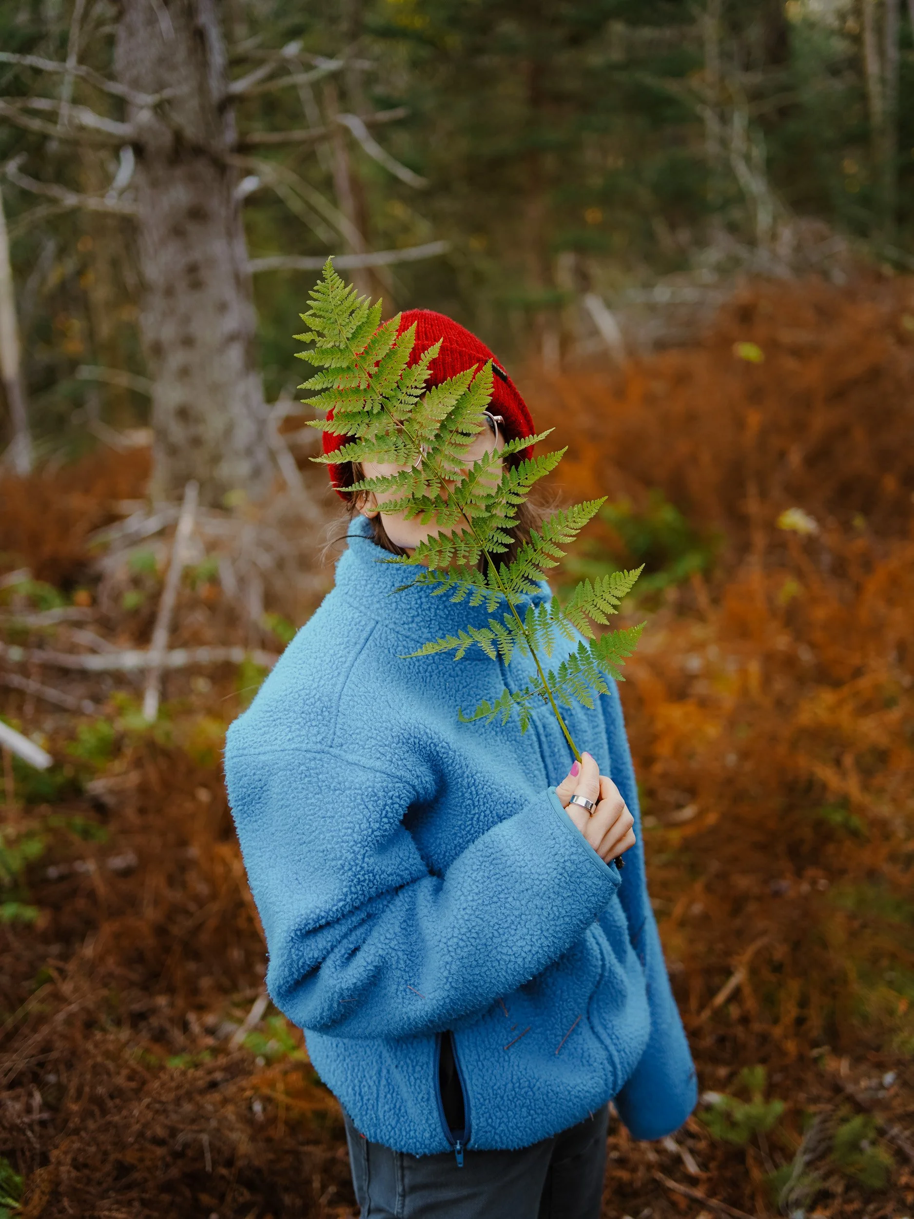 Person wearing a red beanie and a blue fleece jacket holding a fern in front of their face in a forest with brown and green foliage.