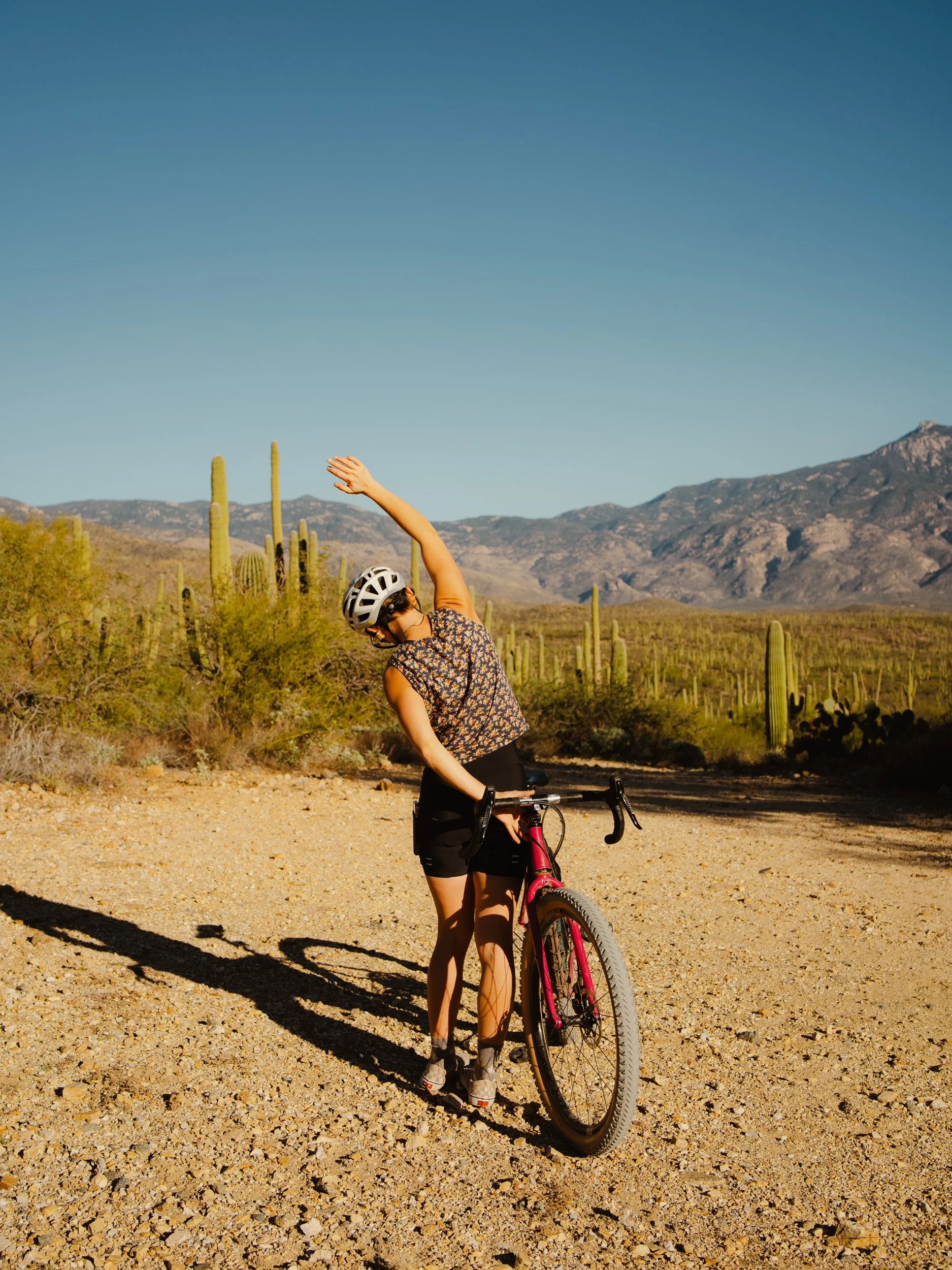 A woman in cycling gear and a helmet stretching in a desert landscape with cacti, mountains, and a clear blue sky.