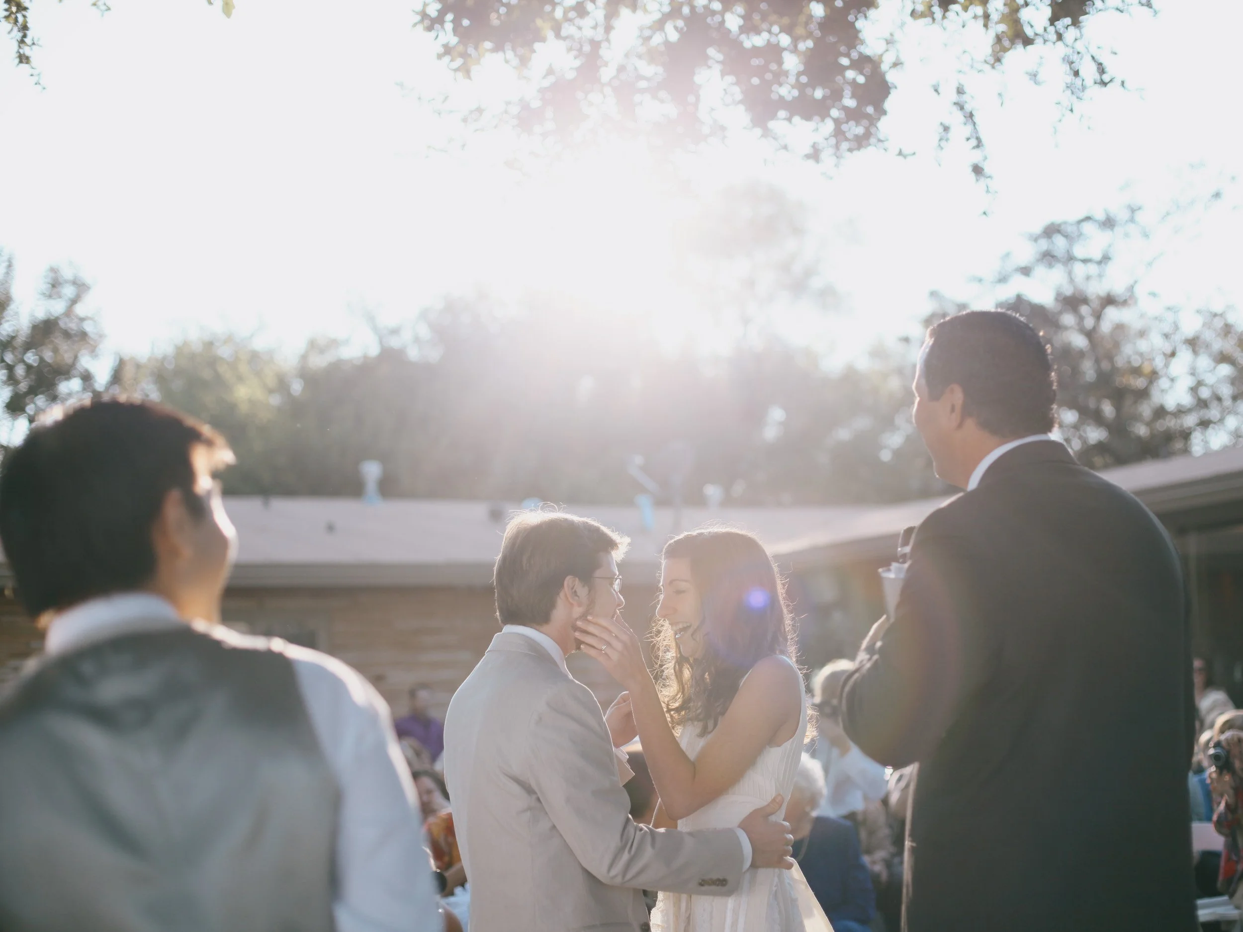 A wedding ceremony outdoors with a bride and groom sharing a moment as they dance surrounded by friends and family, with sunlight filtering through the trees.
