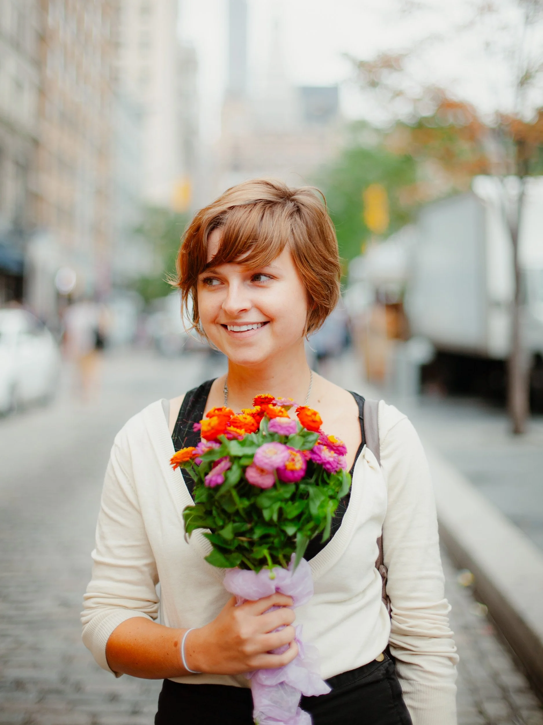 A young woman with short brown hair smiling while holding a colorful bouquet of flowers on a city street.