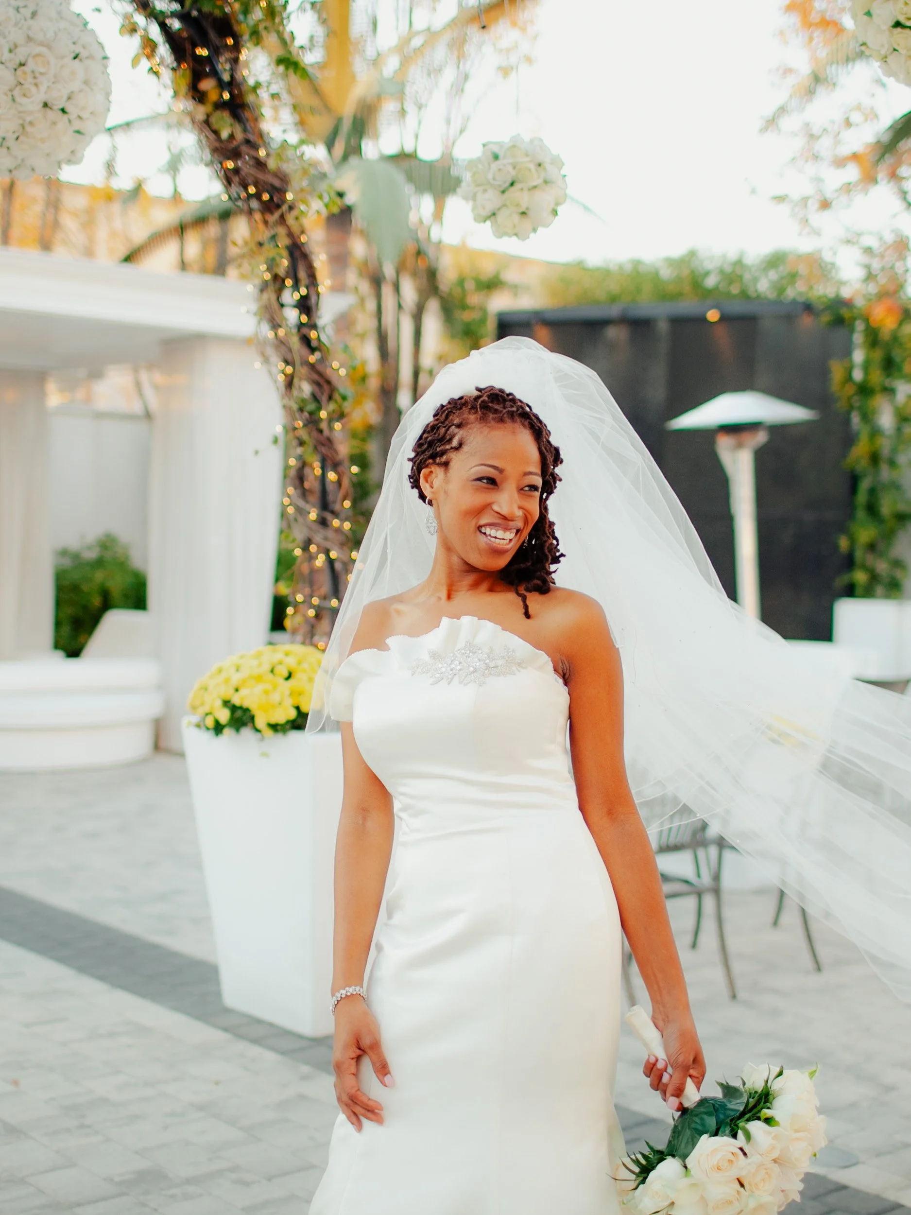 A smiling bride in a white wedding gown holding a bouquet of white roses, outdoors in a decorated venue.