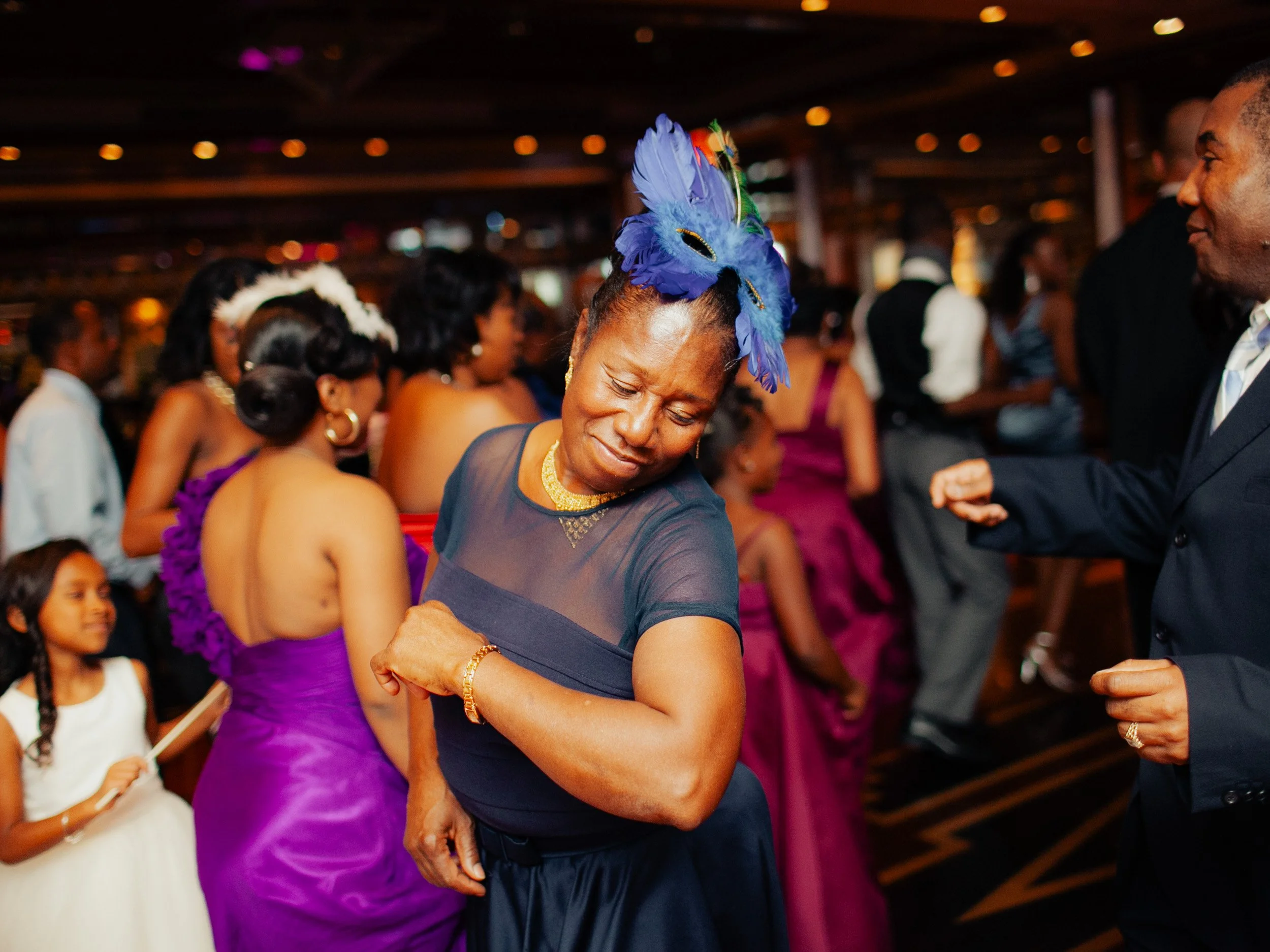 A woman with a blue feathered hairpiece dancing at a party, surrounded by other people in formal attire.