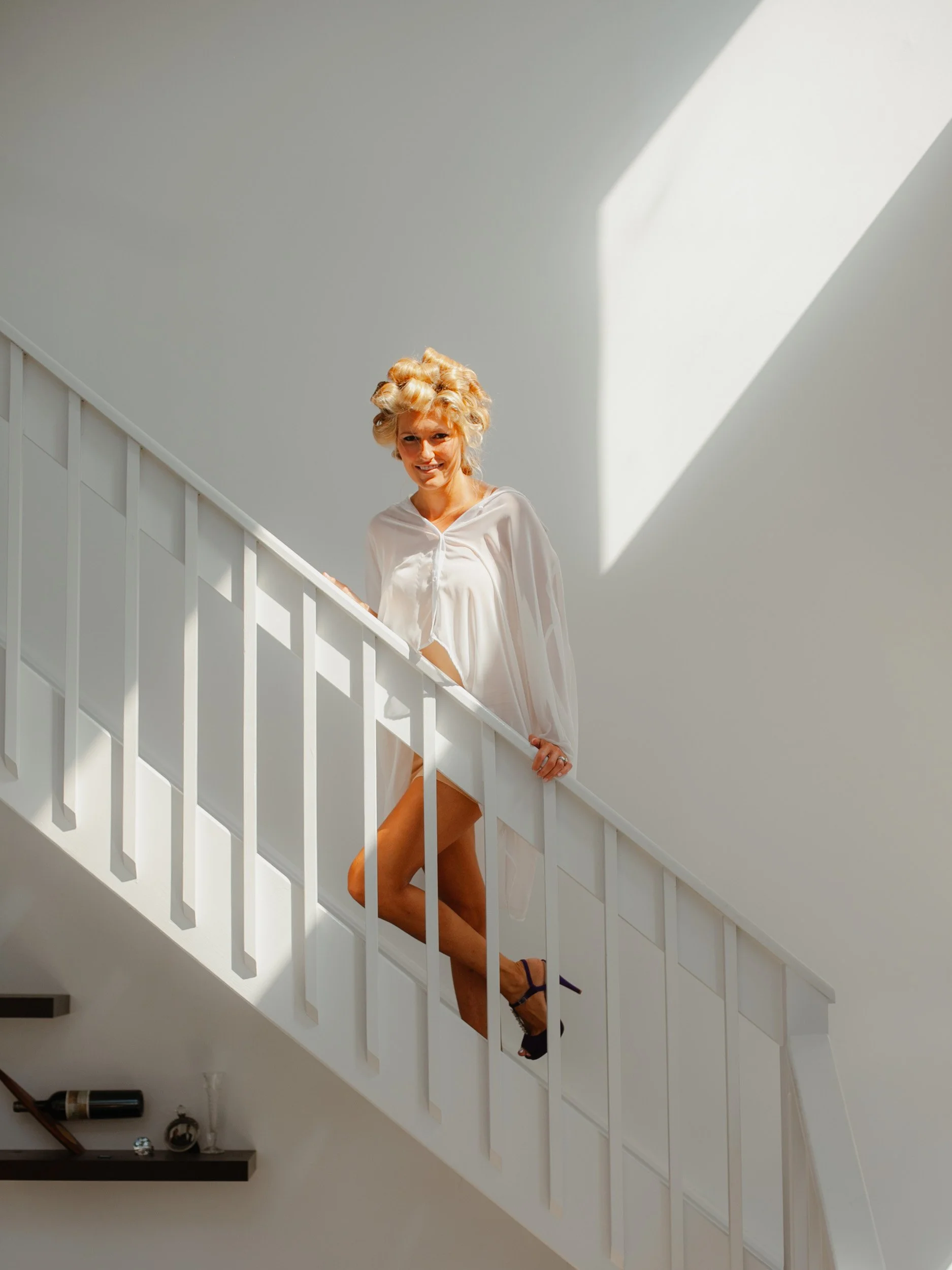 Woman with curly blonde hair wearing a white robe and black high heels on staircase with white railing and wall, sunlight casting a shadow.
