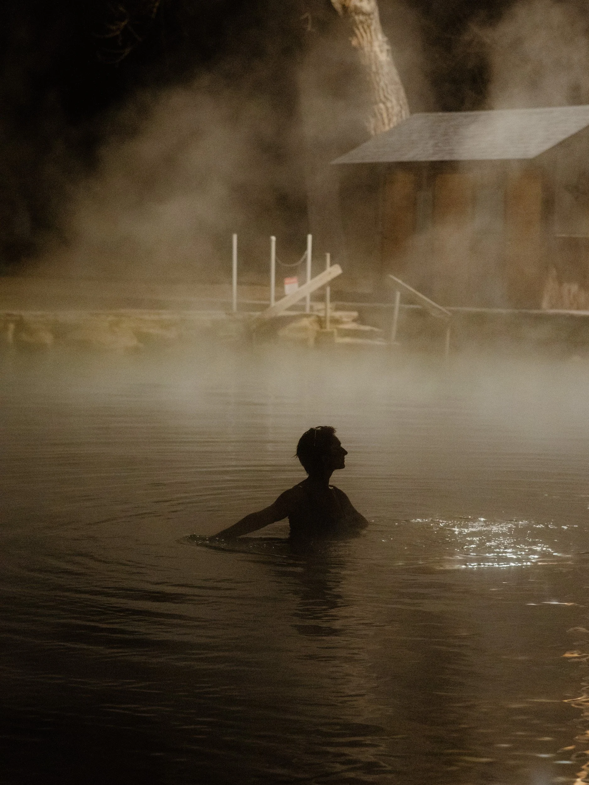 A person is swimming in a hot spring or pool at night, with steam rising around them and a wooden structure in the background.