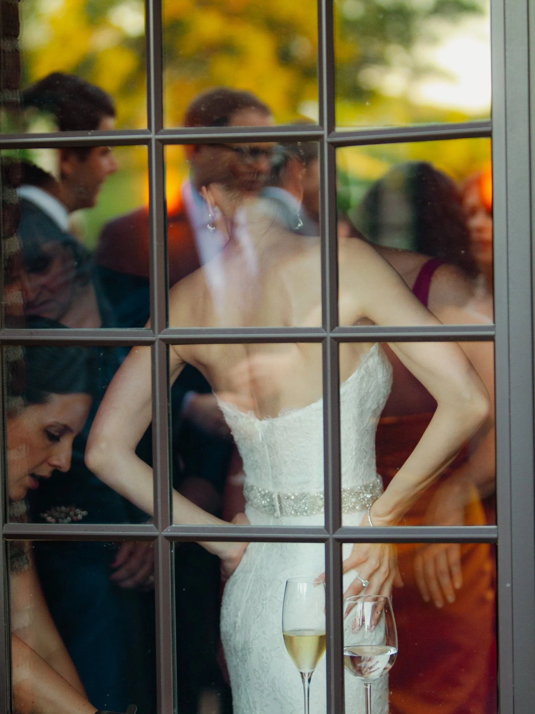 A bride in a strapless wedding gown seen through a window with divided panes, surrounded by wedding guests at an outdoor or indoor event, with glasses of wine on a table in front.