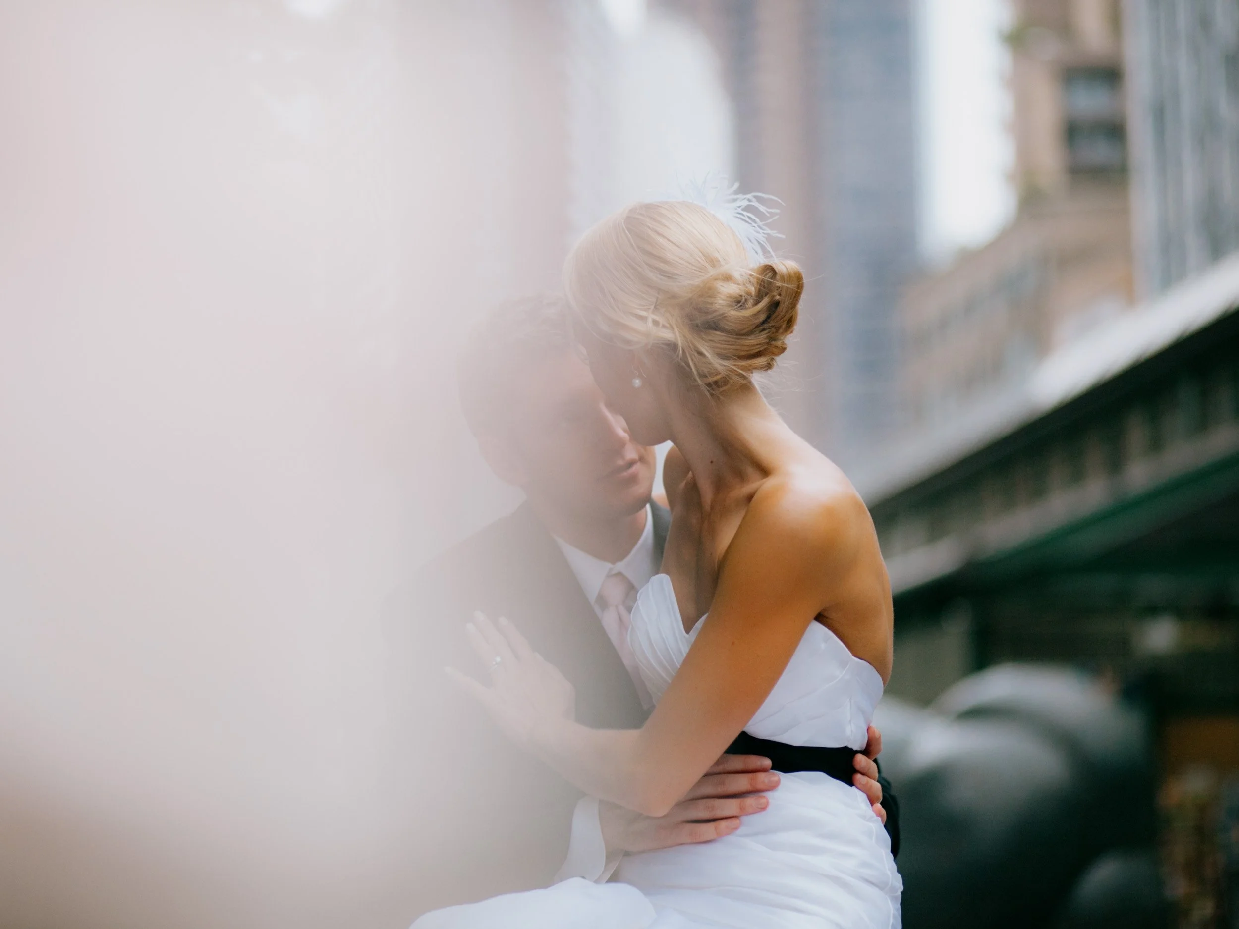 A couple shares an intimate moment on a city street, with the woman in a white strapless dress and the man in a dark suit. They are close, embracing, with blurred city buildings in the background.