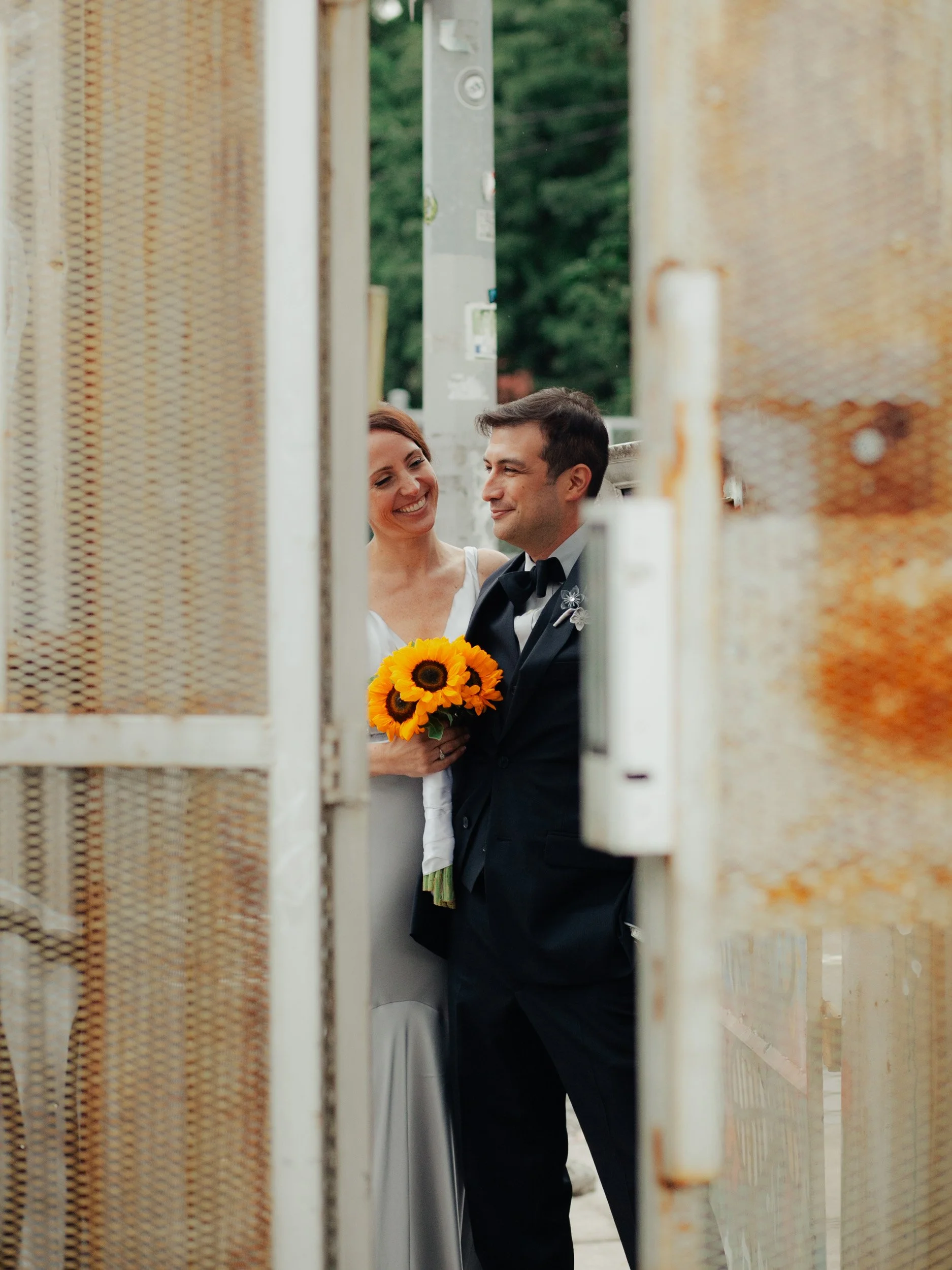 A couple dressed in wedding attire, the woman holding a bouquet of sunflowers, standing close and smiling at each other, seen through a rusty metal door frame.