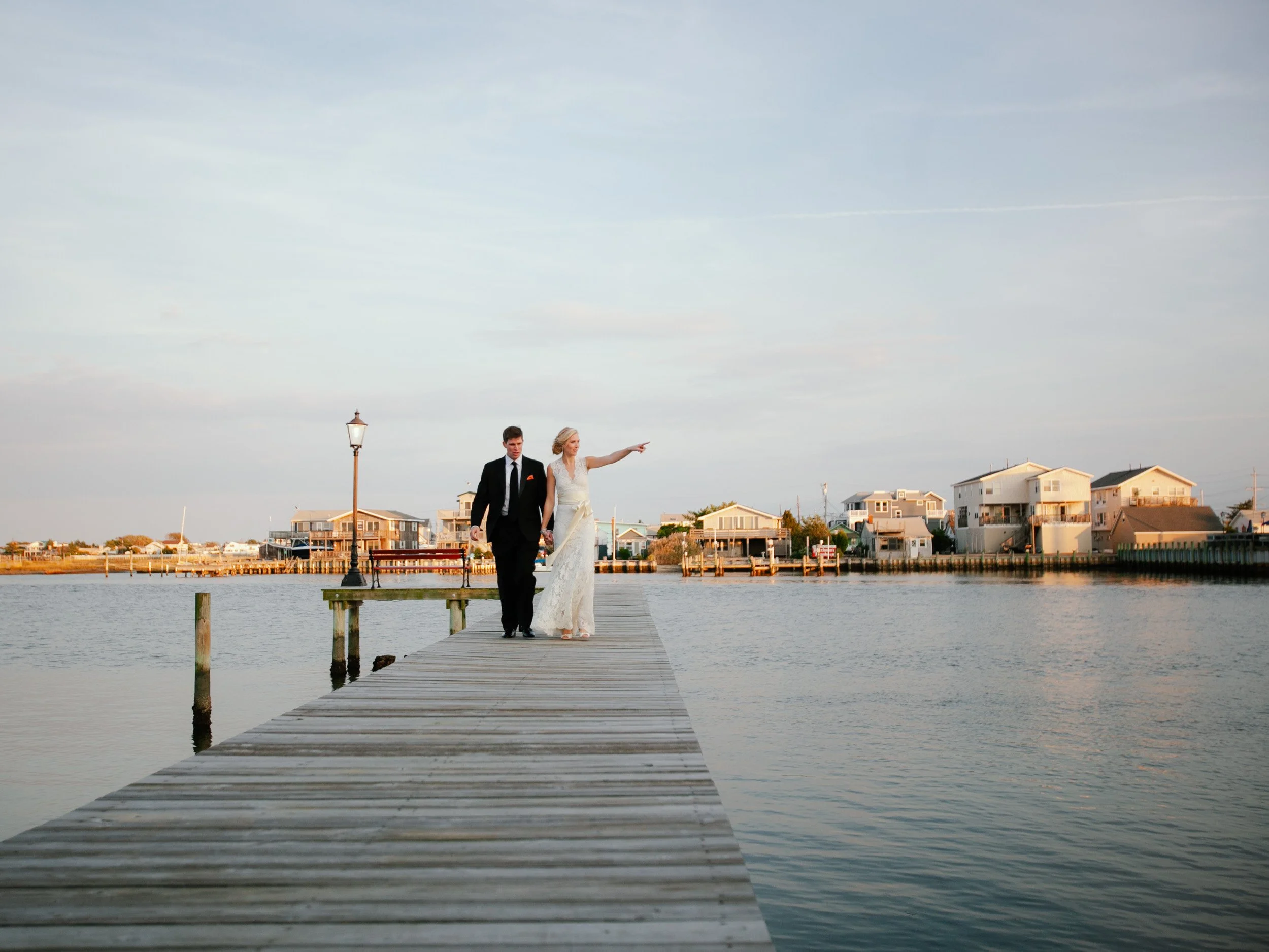 A couple in wedding attire walking on a wooden dock by the water with houses and a lamp post in the background during sunset.