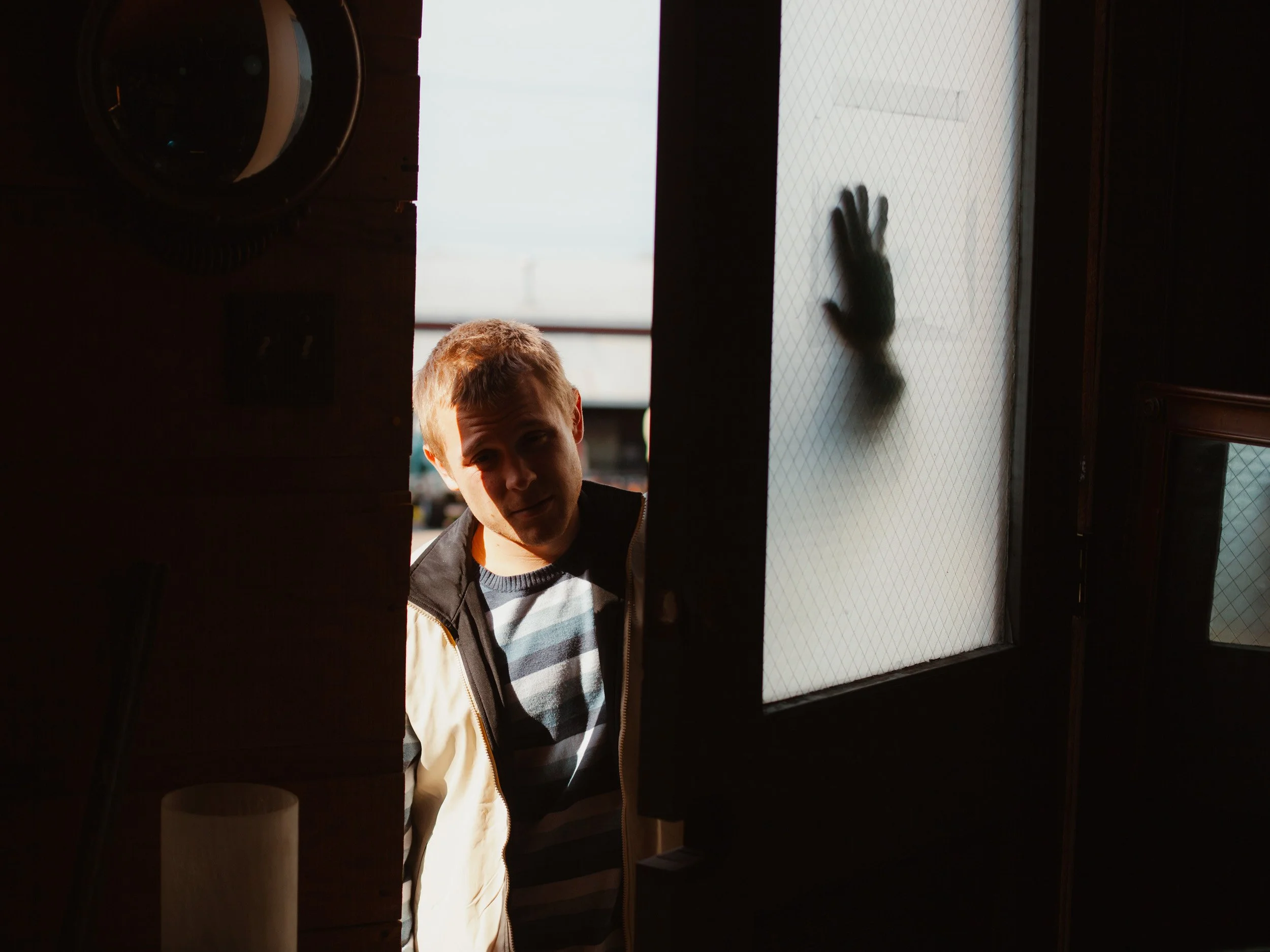 A young man is standing outside a door, with a hand pressing against frosted glass on the door, while sunlight illuminates part of his face.