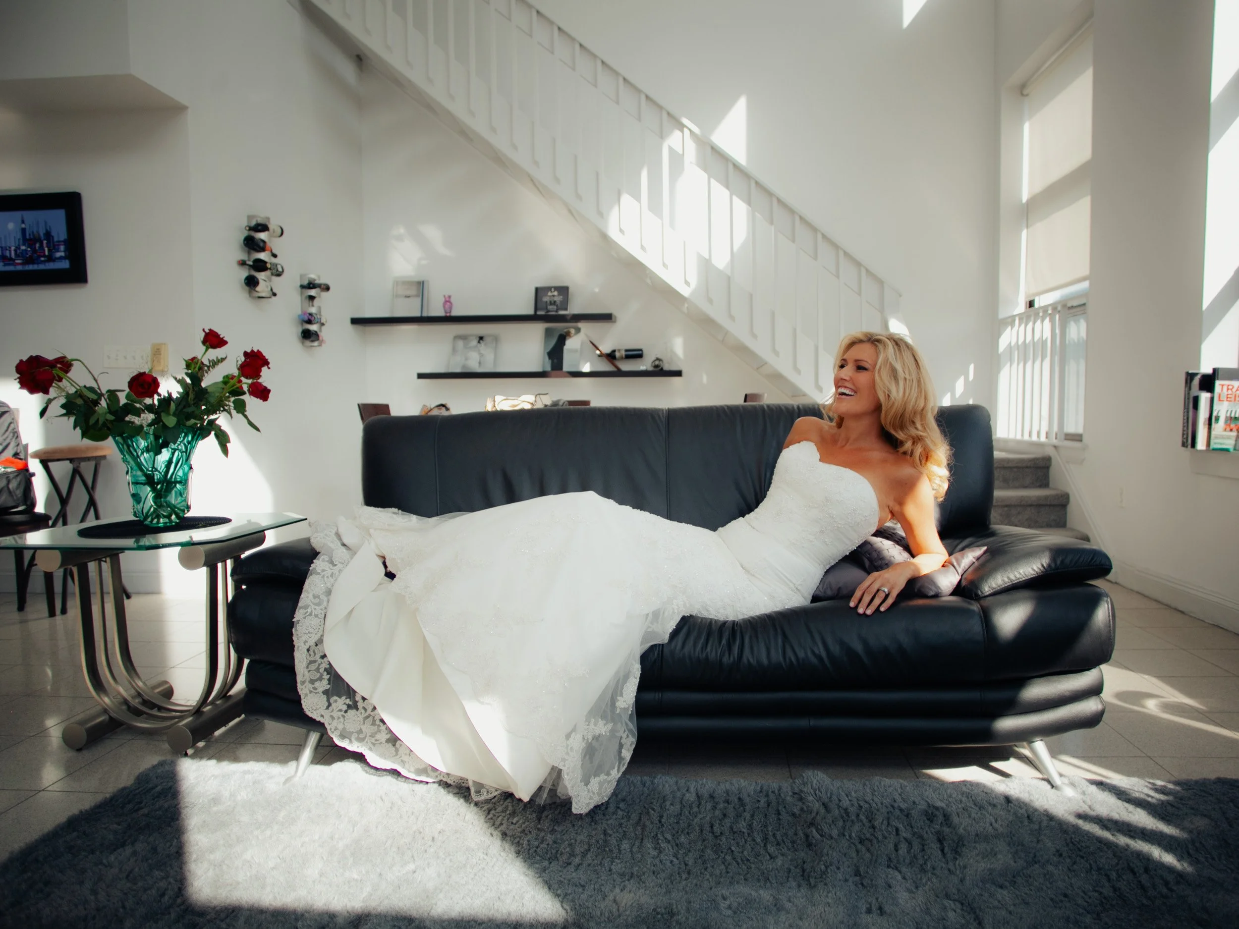 A woman in a white wedding dress lounging on a black leather sofa in a bright living room.