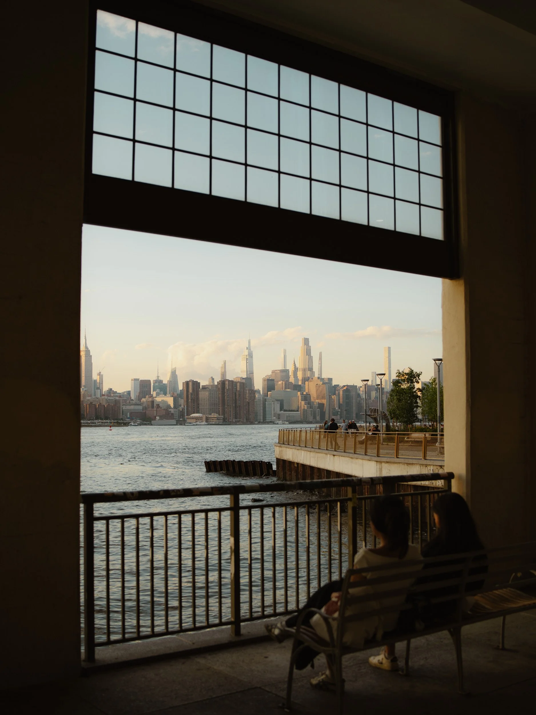 Two women sitting on a bench facing a river with a city skyline in the distance, visible through an open archway with a window above.