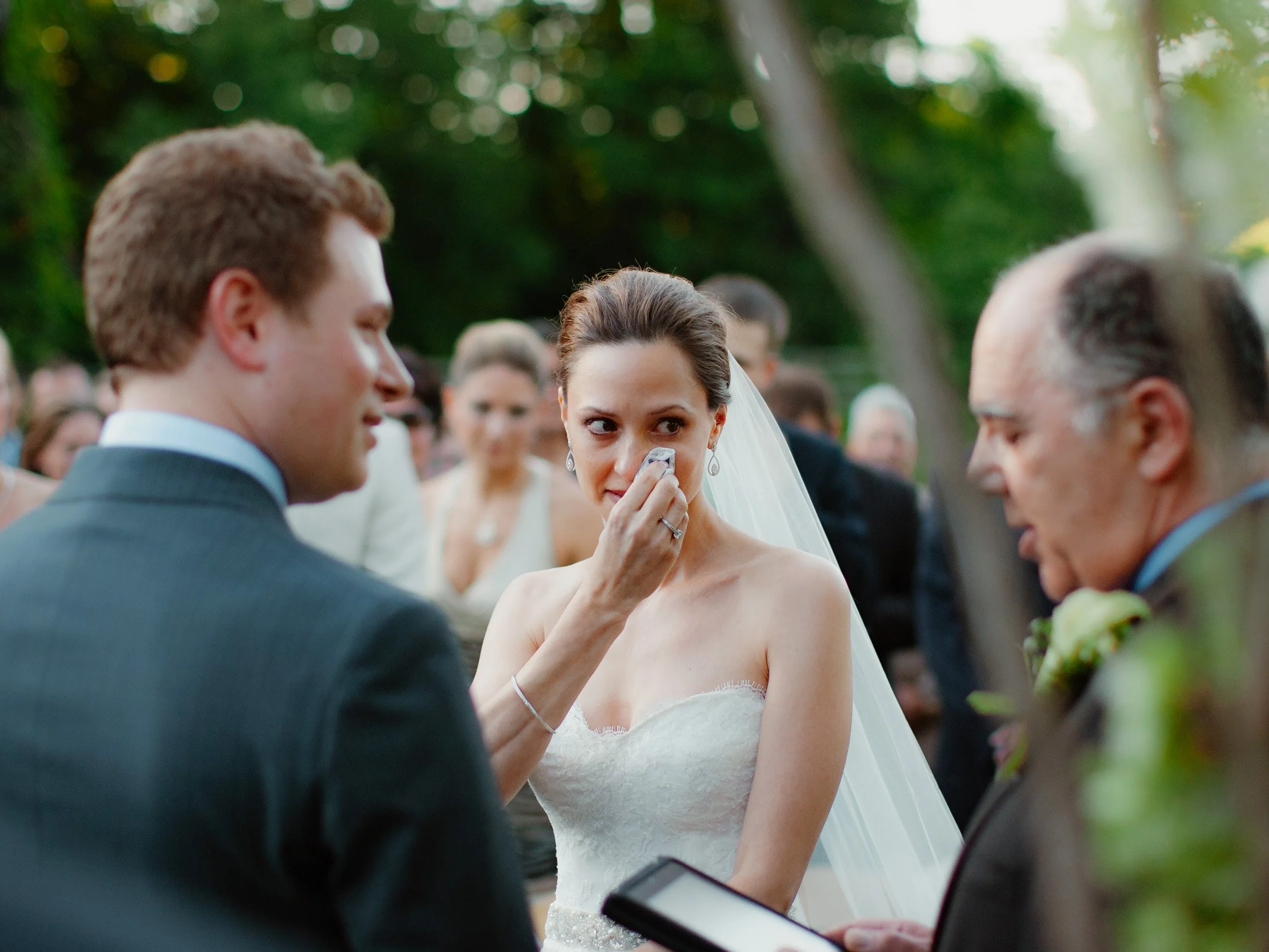 A bride wiping tears during her wedding ceremony outdoors, with a groom and officiant nearby, surrounded by guests and trees.