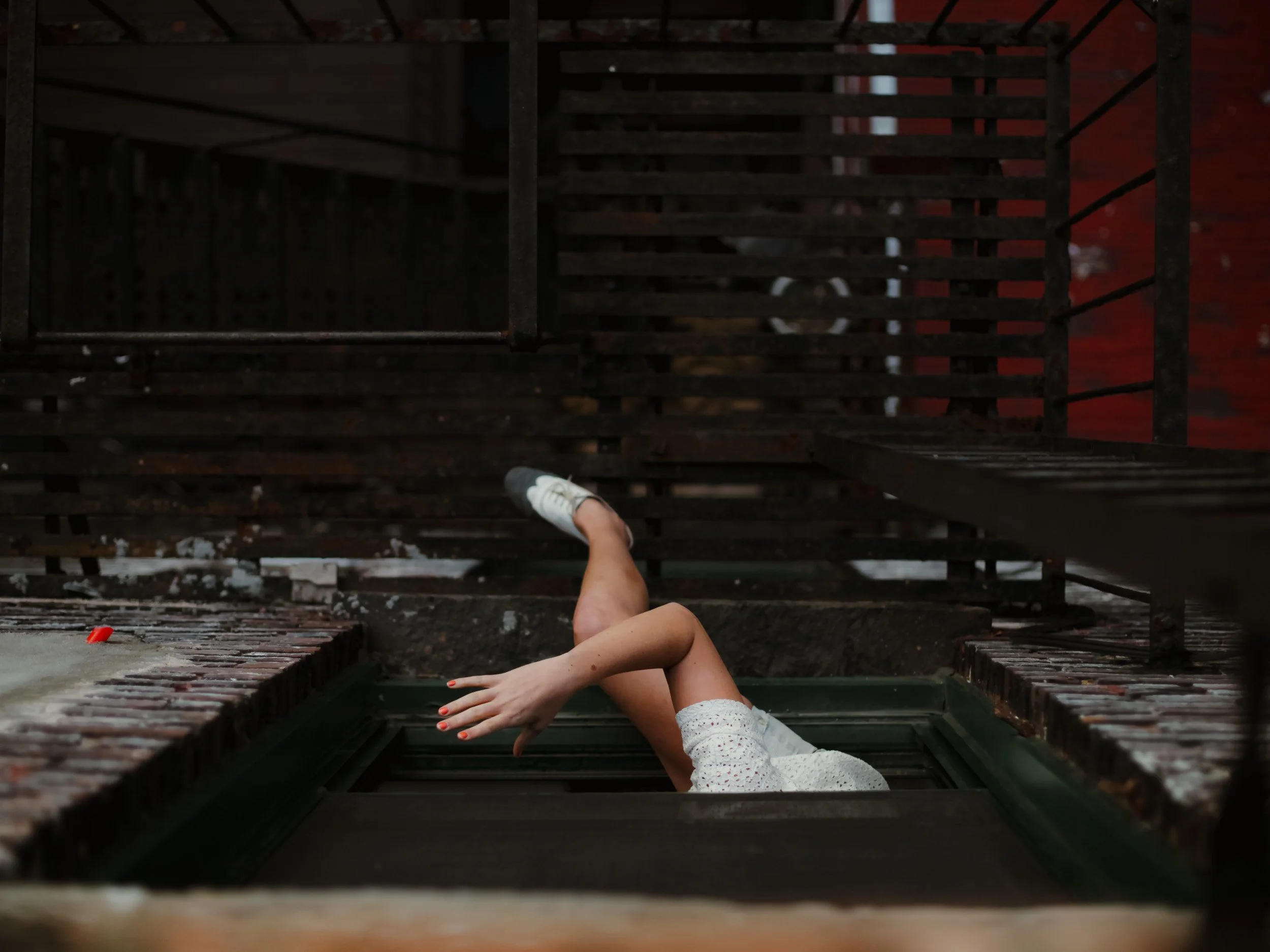A person in a white dress with orange nails and white sneakers is falling headfirst into an open fire escape hatch on a brick building, with their arm extended and legs up in the air.
