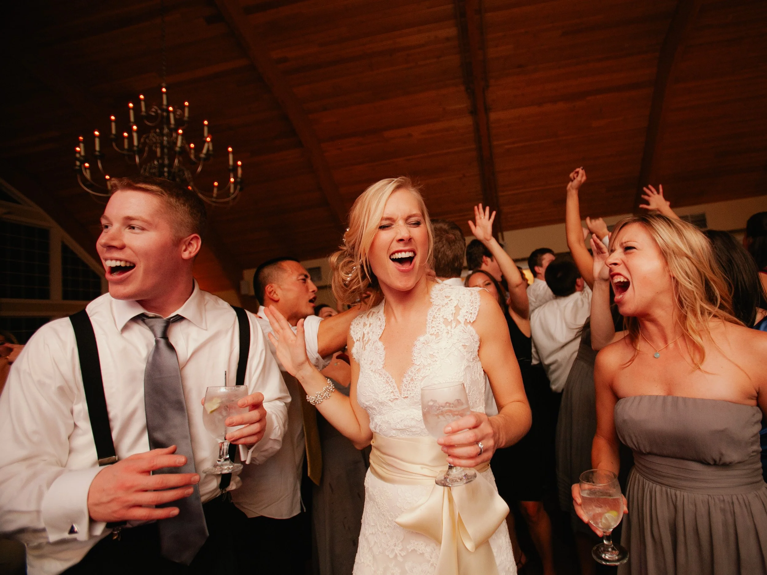 People dancing and celebrating at a wedding reception with a wooden ceiling and chandelier in the background.